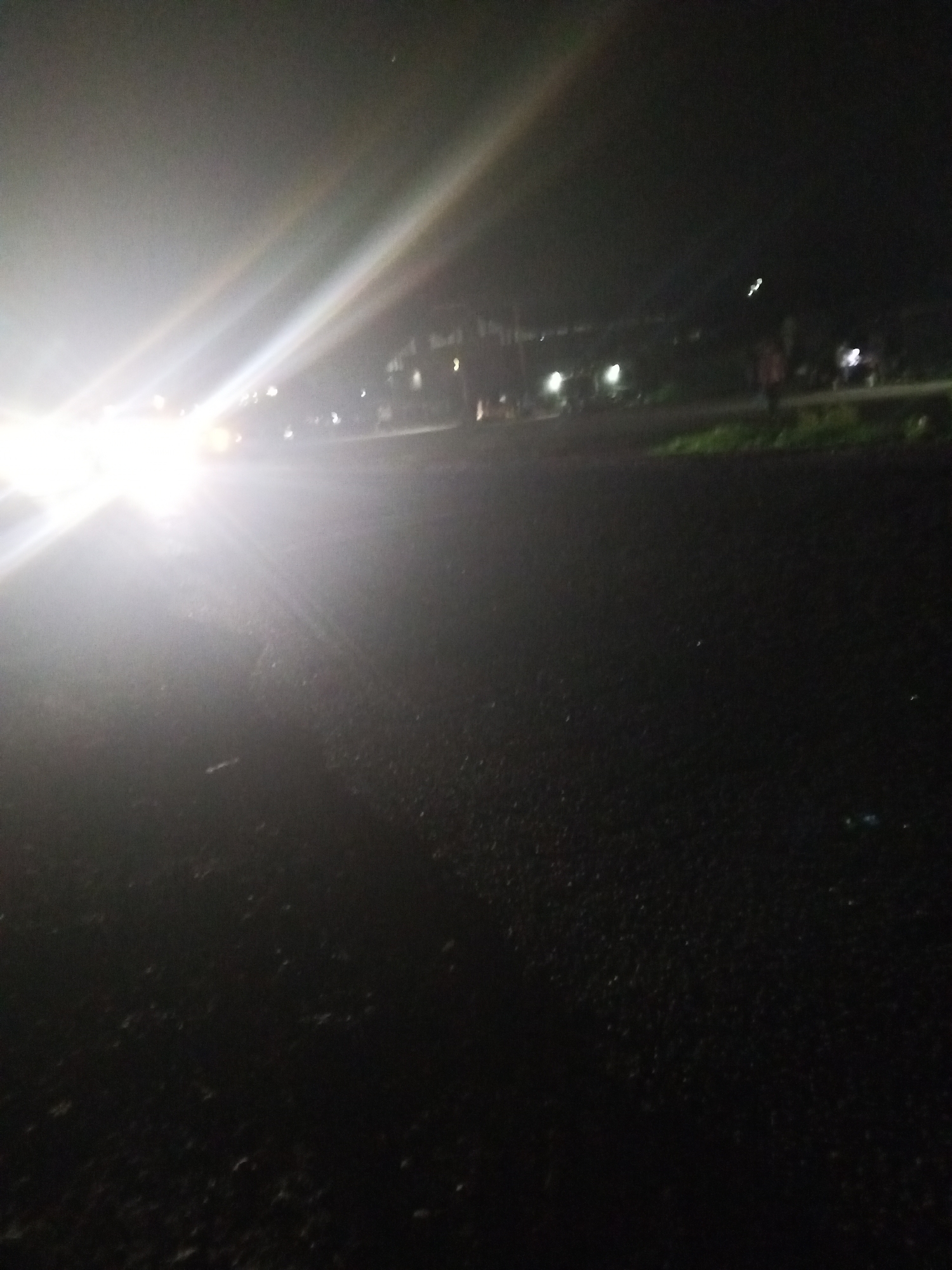 The image depicts a nighttime scene on a road in Lokoja, Nigeria, characterized by heavy light glare from what appears to be oncoming vehicle headlights. The foreground is dominated by a dark asphalt road surface, with scattered debris or small objects visible. In the middle ground and background, the environment is hazy and indistinct due to the low light conditions and glare. There are faint outlines of structures or buildings lining the road, as well as some illuminated points, possibly streetlights or lights from within buildings. The overall impression is that of a street scene at night, possibly during travel, with limited visibility beyond the immediate vicinity due to the intense artificial light sources.