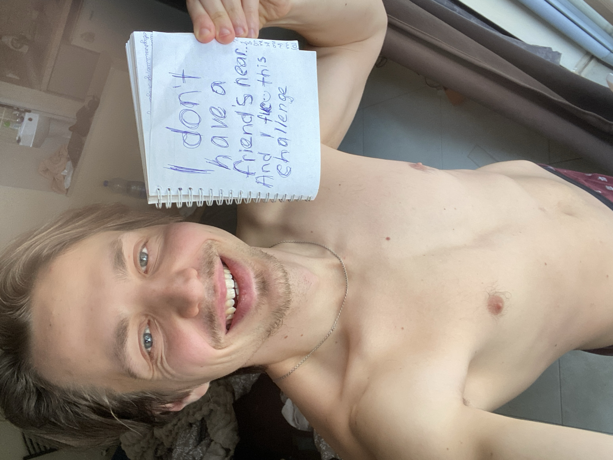 A selfie taken at an oblique angle shows a topless person holding a spiral-bound notebook. The handwritten text on the notebook reads "I don't have a friend's near.. And I like this challenge." The person has blond hair, blue eyes, and a light stubble. A silver chain is visible around their neck. The background includes a partially drawn brown curtain and glimpses of objects on a table and a floor. The location in Šušanj, Montenegro is not discernible from the provided image.