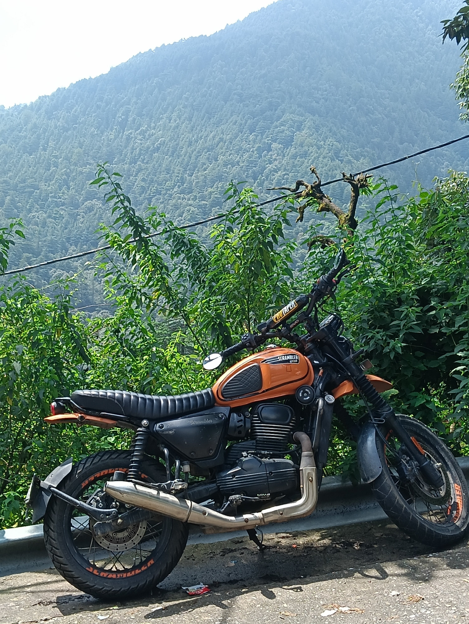An orange and black scrambler motorcycle is parked on the side of a road in Bhagsu, India, with a lush, tree-covered mountainside in the background. The motorcycle is angled towards the left, with its front wheel slightly turned. It has a black ribbed seat, orange accents on the fuel tank, and a silver exhaust pipe. The motorcycle is positioned on a concrete barrier, with a paved road visible in the foreground. Dense green foliage frames the scene, with a power line running horizontally across the upper portion of the image. The sky is bright and overcast.