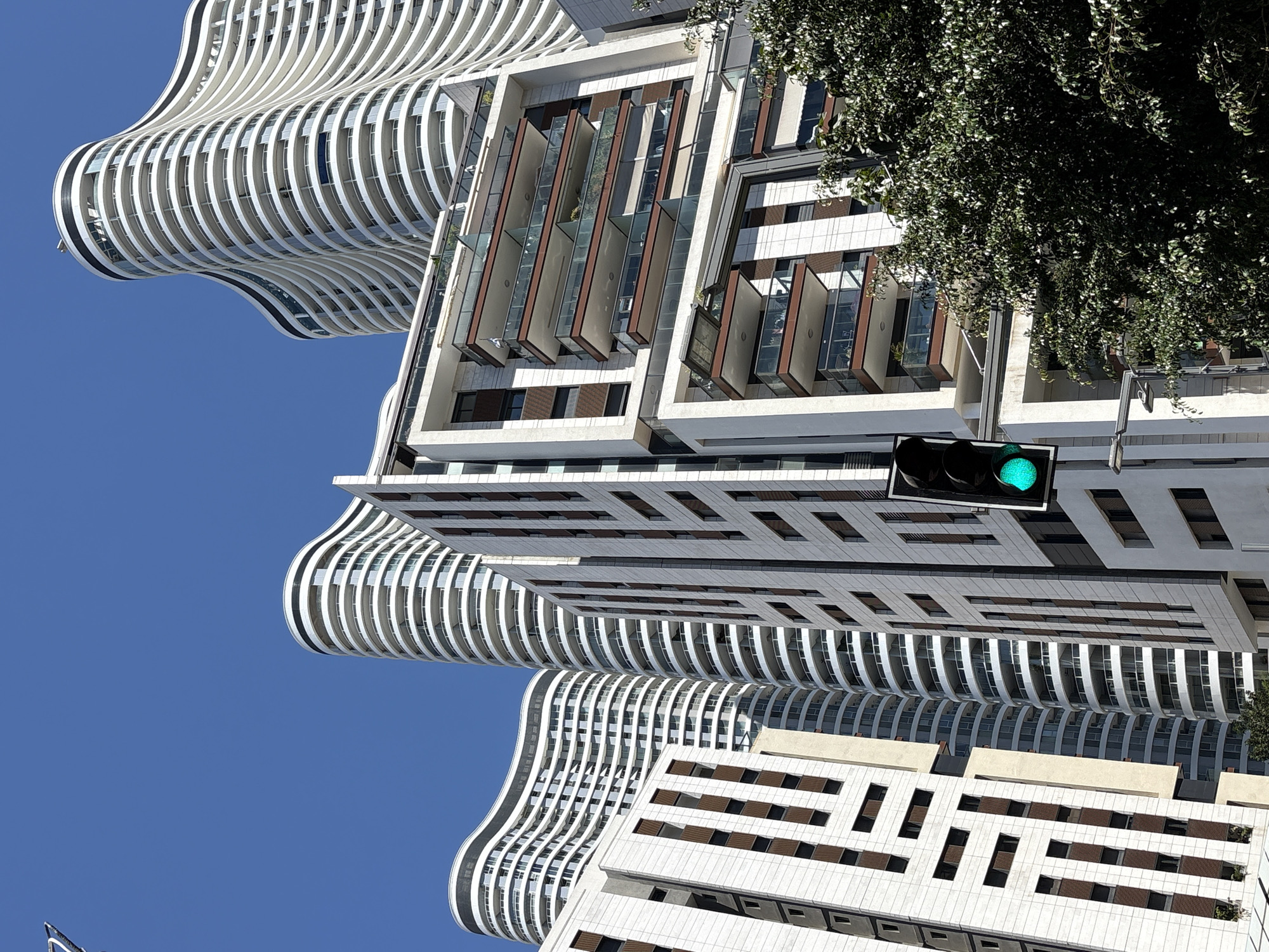 This media file captures a dynamic urban scene in Tel-Aviv, Israel, showcasing modern architectural styles against a clear blue sky. The image is presented with a vertical orientation, causing the buildings to appear rotated and creating an abstract, upward-looking perspective.
The primary subjects are several multi-story buildings, each with distinctive designs. A prominent feature is a building with a wavy, undulating white facade, characterized by numerous horizontal bands that form curved balconies or window outlines. Further to the right, another building exhibits a more angular, multi-layered design with sections of pivoting or recessed windows/balcony enclosures made of glass and reddish-brown panels, set within a white structural frame. Parts of other white buildings with repeating rectangular window patterns are also visible towards the bottom and far right.
A traffic light, displaying a bright green signal, is centrally positioned in the frame. Above it and extending across the upper right portion of the image is a large, dense tree with lush green foliage, partially obscuring the buildings behind it.
The scene takes place on a bright, sunny day, indicated by the vibrant blue sky and strong light casting shadows. No human activity or visible text is present in the image. The unique architectural forms and clear weather contribute to a modern and striking urban landscape.
