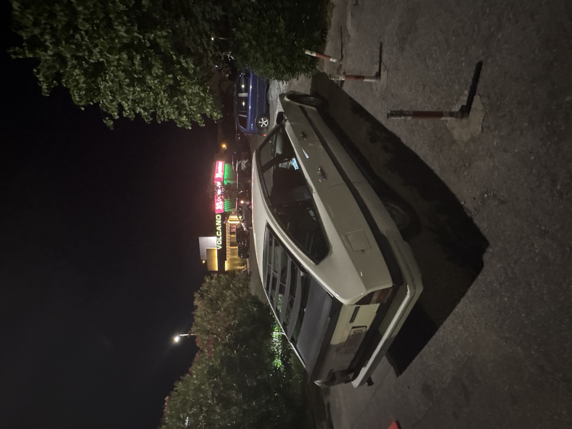 The image depicts a night scene in Podgorica, Montenegro. A white car is parked on the asphalt in the foreground, casting a shadow on the ground. A blue car is parked nearby, partially obscured by trees. In the background is a building with a sign that says "VOLCANO" in illuminated letters. Trees are on either side of the cars. Red and white bollards are along the side of the car. The lighting is dim, with the streetlights illuminating the scene.