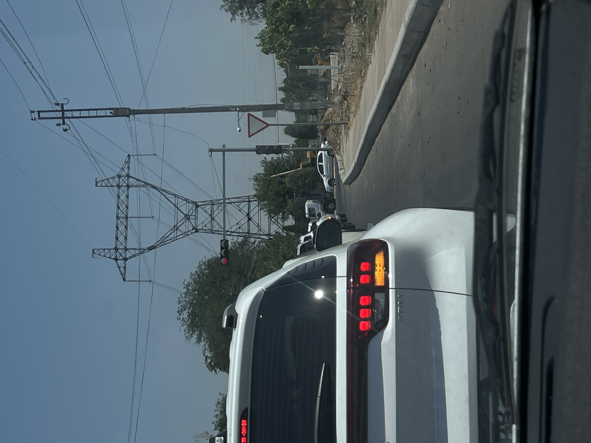 The image captures a traffic scene on a sunny day in Kibray, Uzbekistan. The foreground shows the rear of a white SUV, its brake lights illuminated. In the middle ground, a line of cars is stopped, including a white truck and a white sedan. The background features a large electricity pylon with overhead wires, a traffic light, and a yield sign. Trees and construction activity, including a yellow excavator, are visible along the roadside. The scene appears to be a typical urban traffic situation with infrastructure and natural elements.