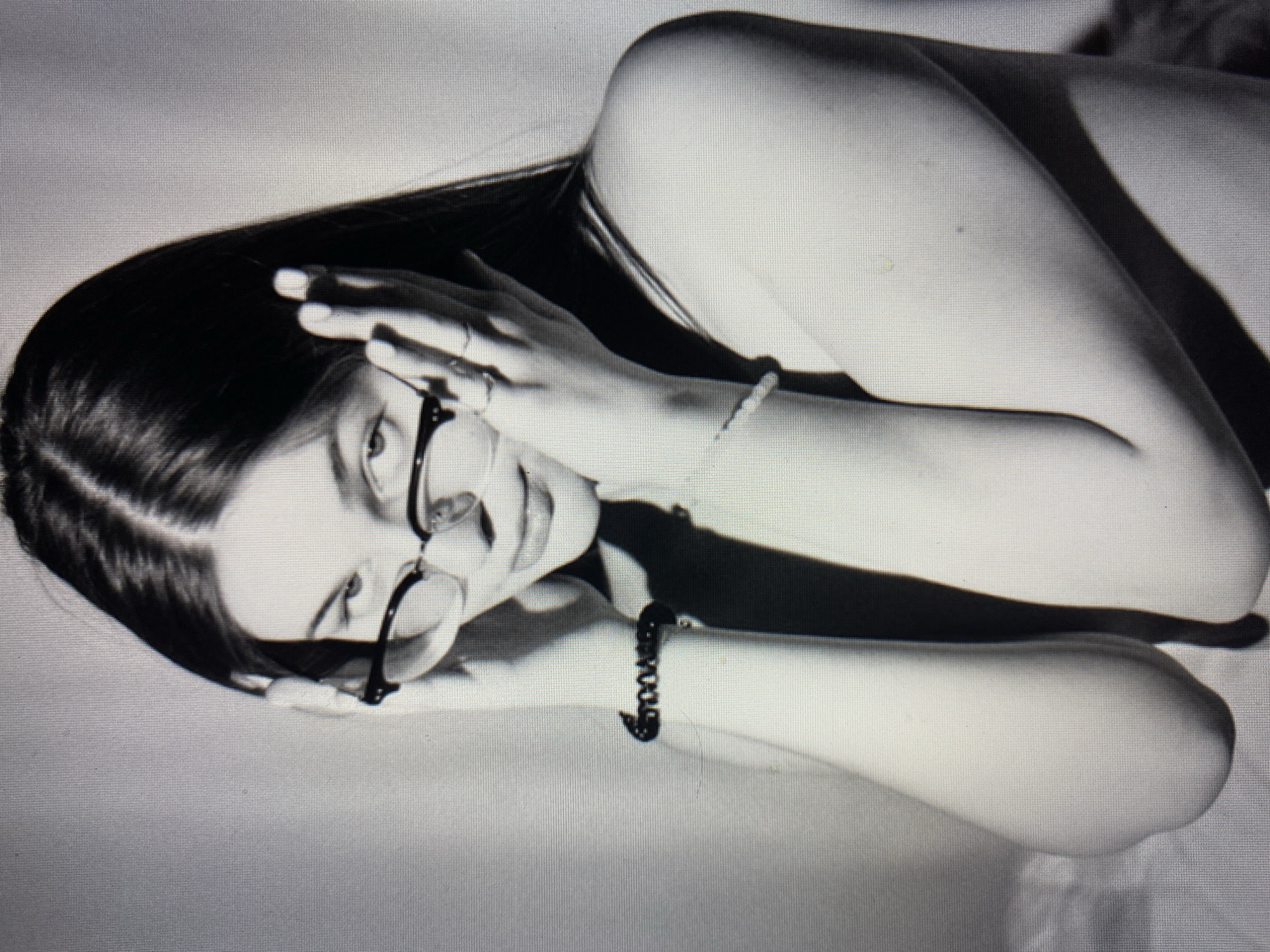 A black and white, medium shot photograph shows a young woman wearing glasses and looking directly at the camera. She has her hands on either side of her head, with her fingers touching her temples. Her hair is dark and long, falling over her shoulders. She is wearing a sleeveless top, and her left arm is visible, adorned with a bracelet. The background is a plain, textured wall. The lighting is dramatic, casting shadows on the woman's face and body.