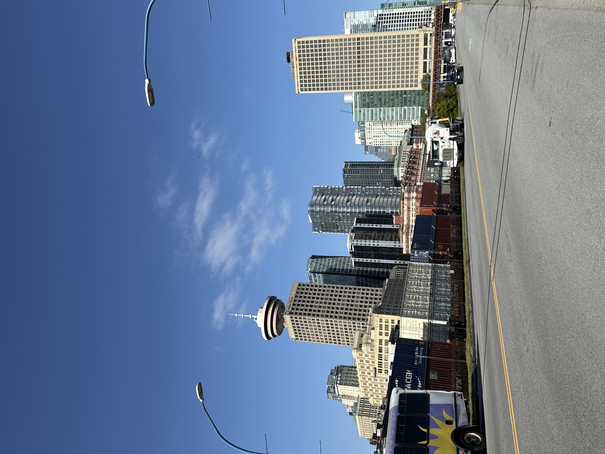 This media file captures a vibrant urban scene in Vancouver, Canada, under a clear blue sky with sparse white clouds. The image is oriented sideways but depicts a street running along a city skyline, featuring numerous high-rise buildings and modern skyscrapers.
Prominently featured is the Vancouver Lookout (Harbour Centre) with its distinctive saucer-shaped observation deck and spire, confirming the location. Several other buildings, varying in architectural style from older brick structures to contemporary glass towers, form a dense cityscape.
Along the right side of the image, an asphalt road with a double yellow line indicates a thoroughfare. Parked or driving on and alongside this road are various vehicles, including several large semi-trucks, one white truck clearly visible with its cab. Numerous colorful shipping containers, likely part of a port or industrial area, are stacked along the road, separating it from some of the buildings. One blue container has the text "ACGM" visible. A colorful bus, primarily purple and yellow with a sun-like graphic, is also parked, displaying partial text on its side: "... on the things you can do".
Two modern streetlights with long, curved blue poles extend into the clear sky from the left side of the frame. The bright, direct sunlight suggests it is midday or early afternoon. The overall impression is a bustling urban and industrial landscape under sunny weather.