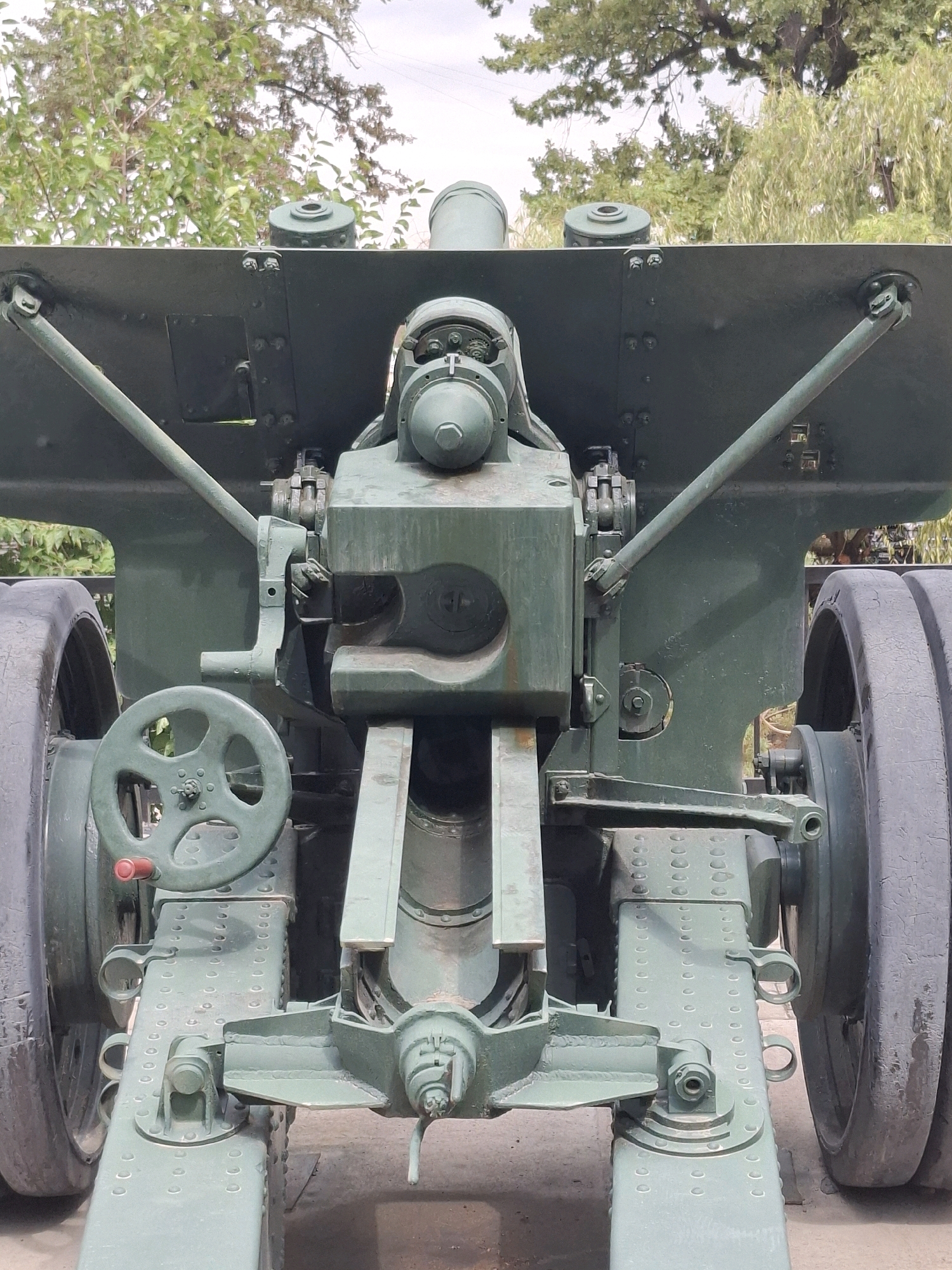 The image shows a close-up, frontal view of a large, green artillery gun. The gun is stationary, resting on its wheels and part of its carriage. The carriage features a large, green steering wheel on the left side, and various mechanical components, including levers and gears, are visible. The gun is positioned outdoors, with trees and foliage visible in the background under a bright, overcast sky. The scene appears to be a static display, possibly in a park or museum, given the lack of human activity and the well-maintained appearance of the artillery piece. Without further information or context, the specific location within Almaty, Kazakhstan, cannot be determined from the visual elements alone.