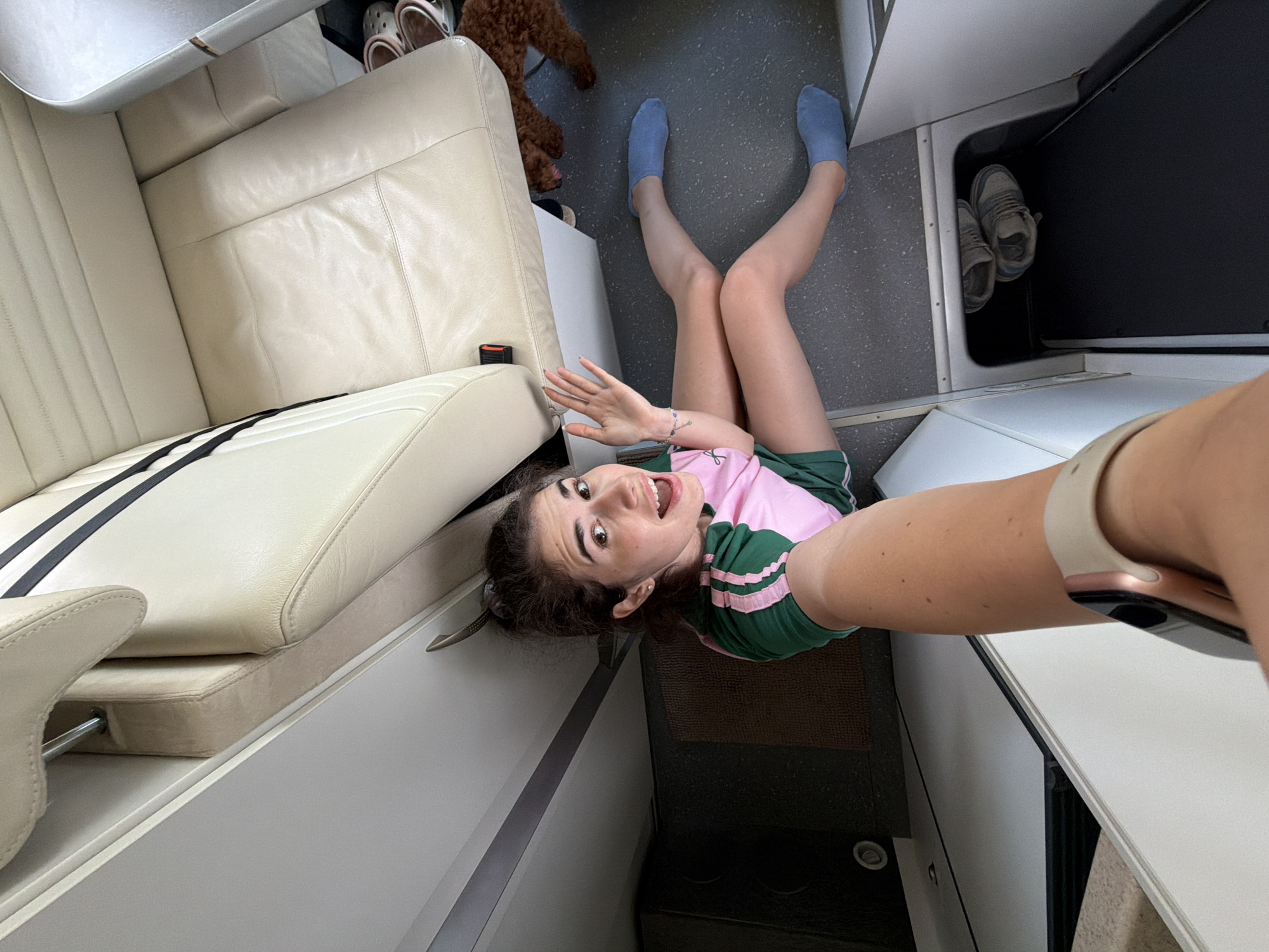 A young woman with dark hair is sitting on the floor of what appears to be a camper van or recreational vehicle, taking a selfie. She is wearing a pink and green t-shirt and blue socks. Her legs are extended in front of her, and she is smiling and waving at the camera. A small brown poodle is visible in the background, partially obscured by the woman's legs and the seat. The interior of the vehicle is clean and organized, with light-colored seats and white cabinets. The floor is a speckled grey material. There is a shoe storage compartment visible below the seats containing a pair of sneakers. The scene appears to be indoors, possibly in the city of Nea Moudania, Greece, based on the context provided.