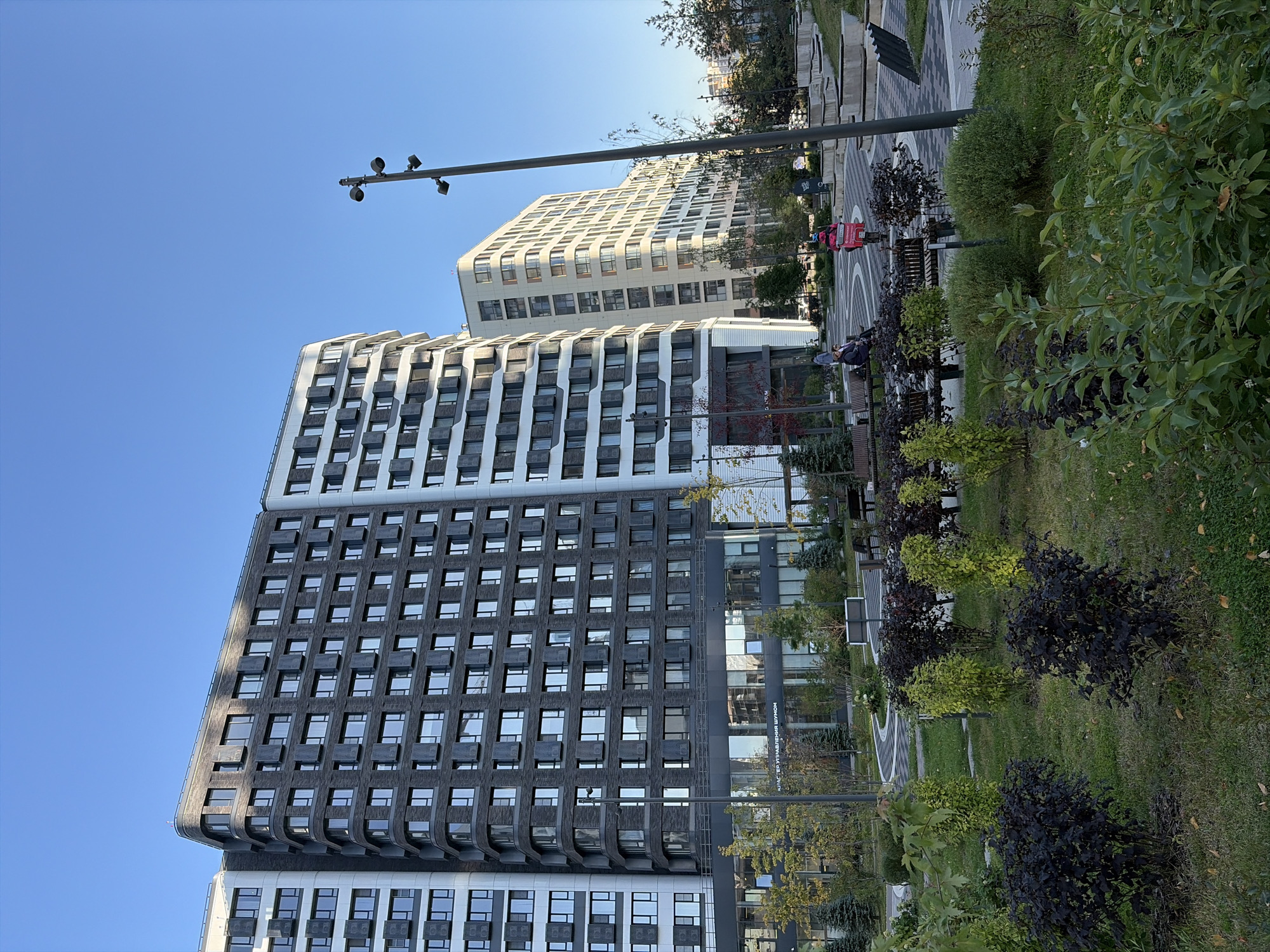 The image depicts a modern residential complex in Moscow, Russia, characterized by its contemporary architecture. In the foreground, a well-maintained park area features lush green grass and various shrubs, including dark purple and lime green varieties. Several park benches are visible, with two individuals seated on one of them. Further into the park, a person on a pink scooter with a delivery box is seen riding through a paved area with geometric patterns.
Dominating the background are two tall, multi-story buildings. The building on the left is larger, featuring a dark gray facade with a repeating pattern of windows and dark gray exterior elements. Its curved corner suggests a modern design. To its right, a lighter-colored building stands, also with numerous windows and a slightly curved profile. The sky above is a clear, bright blue, indicating fair weather. The scene appears to be a residential courtyard or public space within a modern urban development.