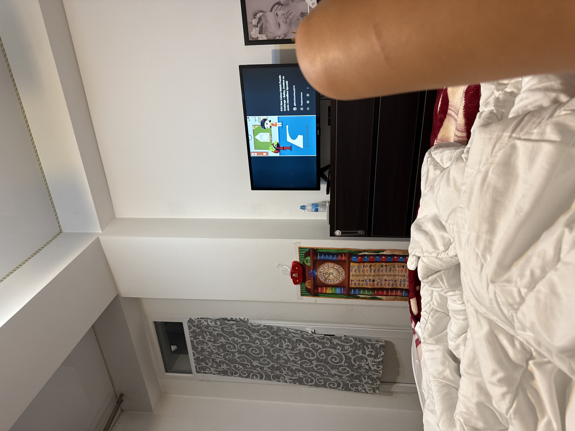 The image depicts a bedroom scene, likely in Chișinău, Moldova, as indicated by the presence of Cyrillic script on the educational chart on the wall. The room appears to be furnished with a bed in the foreground, covered in white bedding. To the right, a dark wood dresser is visible, topped with a television displaying a children's game or animation. A framed photograph of a baby is positioned to the right of the television. On the wall to the left, a tall, narrow window is partially covered by a grey patterned curtain. Adjacent to the window, an educational chart with a clock and Cyrillic characters hangs on the wall. The overall impression is that of a domestic living space.