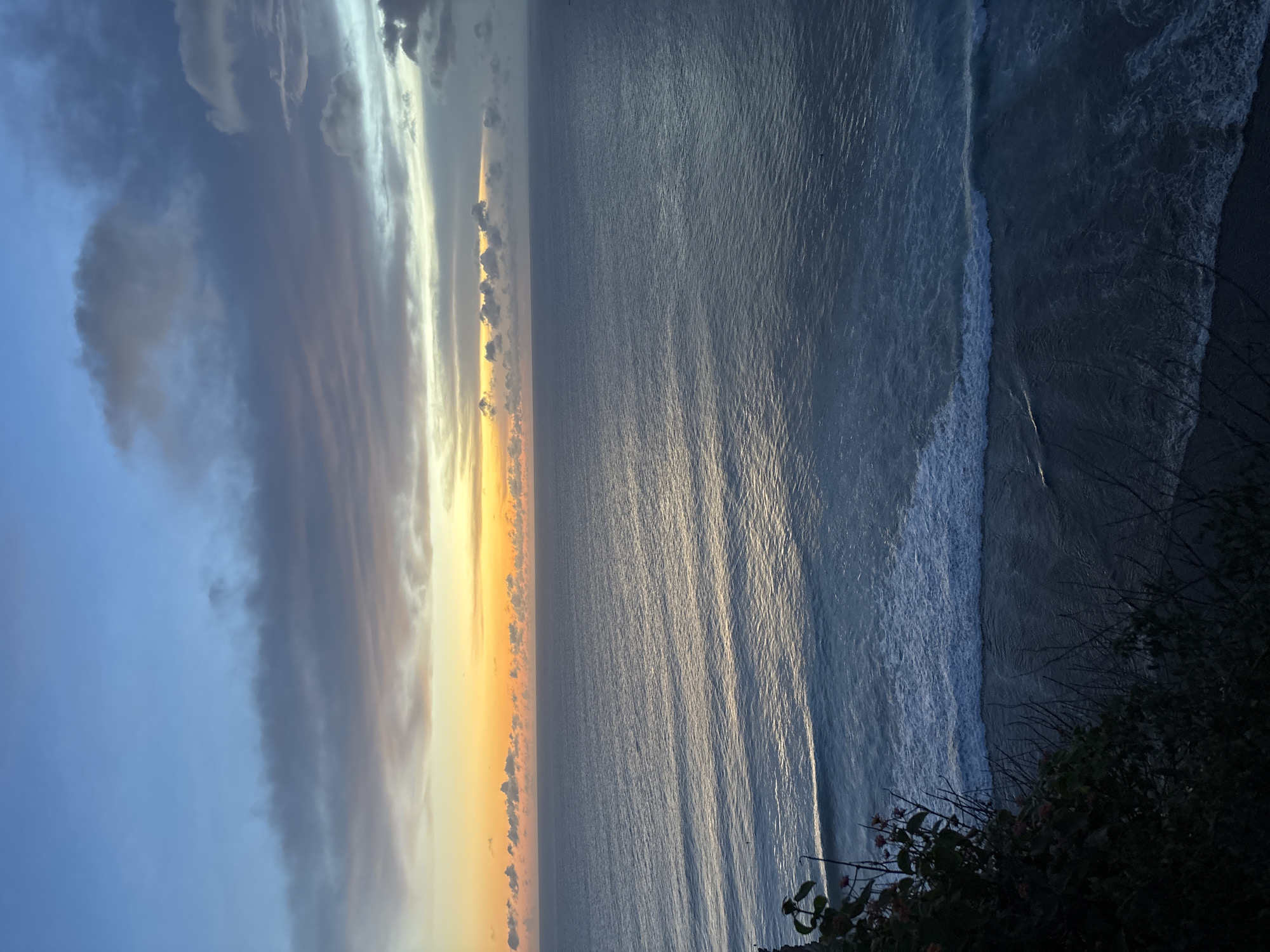 The image shows a vertical view of the ocean during a sunset in Uluwatu, Indonesia. The ocean stretches from the middle of the frame to the right edge, exhibiting subtle wave patterns. The sky, on the left side, is painted with shades of blue, gray, and streaks of orange sunlight piercing through the clouds. A thin line of vibrant orange light separates the clouds from the ocean. The foreground is slightly obscured by foliage in the bottom right corner.