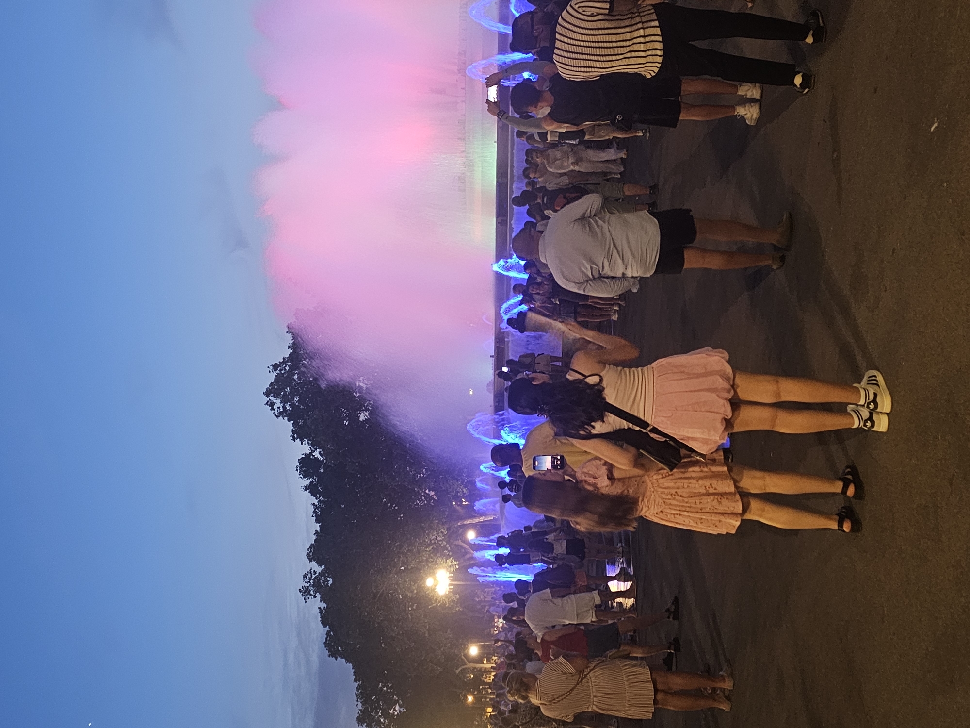A group of people are gathered in front of a colorful light and water show in Barcelona, Spain. The event appears to be taking place at dusk, as the sky is a deep blue with hints of pink and purple from the light display. The water jets are illuminated with vibrant colors, creating a mesmerizing spectacle.
The crowd consists of individuals of various ages, some standing and watching the show, while others are taking pictures or videos with their phones. The people are dressed in casual summer attire, suggesting warm weather. Among the crowd, some individuals are holding up their phones to capture the moment. Two women in the foreground are wearing dresses, and one is taking a photo of the fountain.
The scene is set on a paved area, possibly a plaza or a promenade, with trees and streetlights visible in the background. The overall atmosphere seems festive and lively, with a sense of shared enjoyment of the visual performance. There is no visible text in the image.