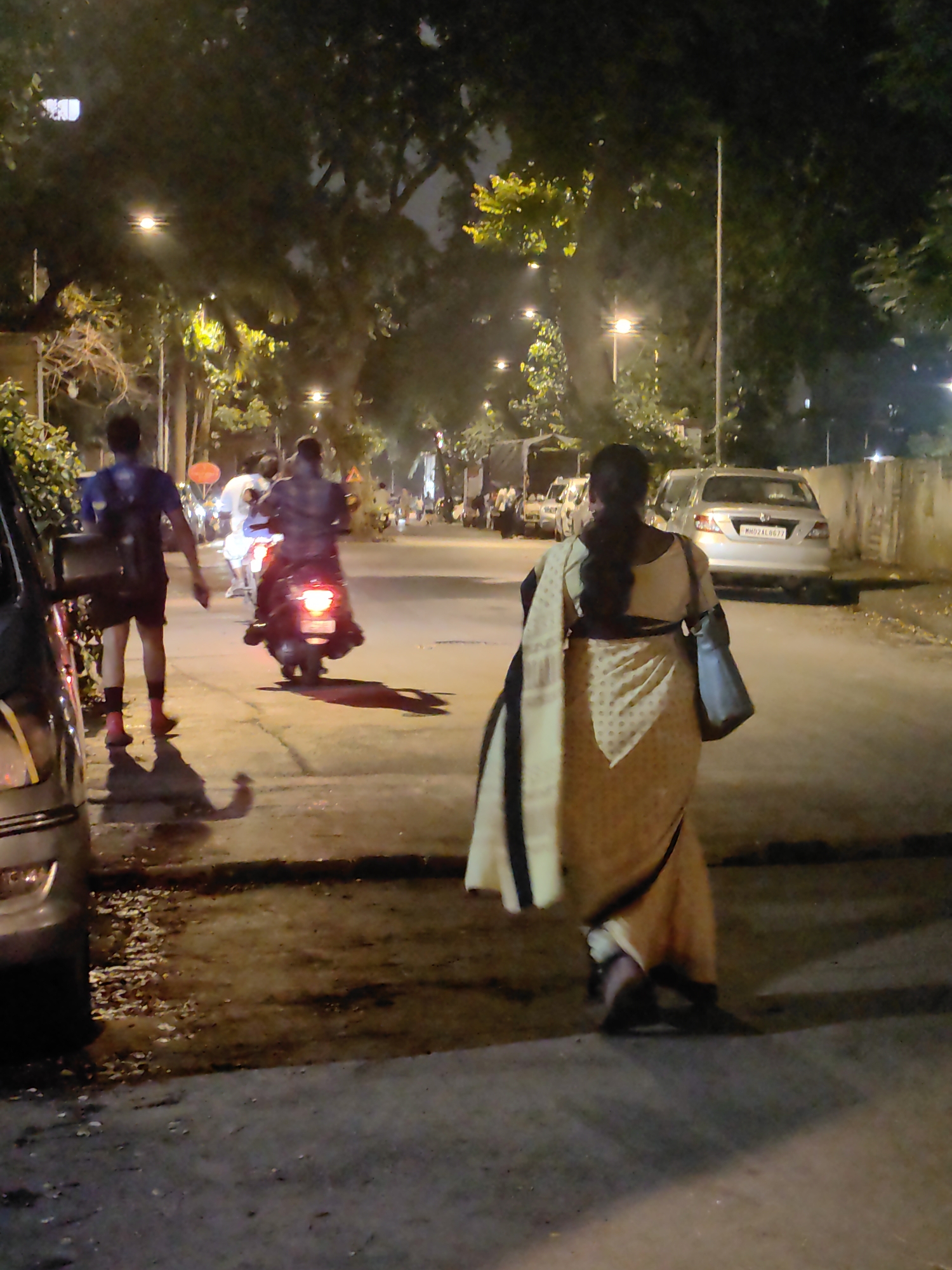 The image depicts a nighttime street scene in Mumbai, India. A woman wearing a sari is walking away from the camera, her sari a light yellow with a patterned border. She carries a dark blue bag over her shoulder. Behind her, a man on a scooter with its brake lights illuminated rides down the street. Another man, wearing a dark blue shirt and shorts, walks on the sidewalk in the foreground, his back to the camera. His shadow is cast onto the street. The street is lined with trees and illuminated by streetlights. Several cars are parked or driving on the road. The ground appears to be a mix of asphalt and concrete, with some debris scattered about. The overall impression is a typical urban evening in India.