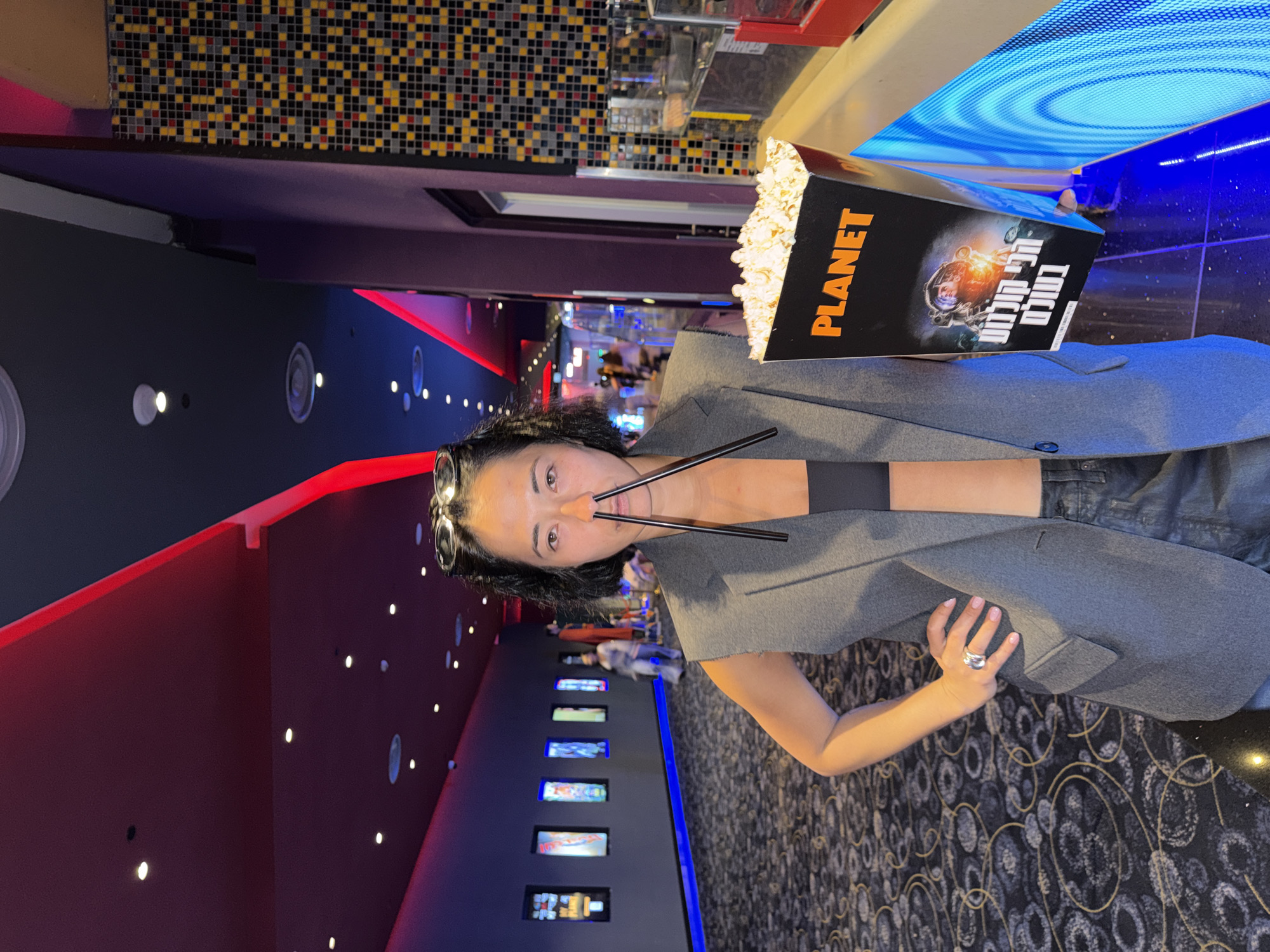 The image shows a woman in a movie theater. She is holding a large box of popcorn with "PLANET" and Hebrew text printed on it. Her face is partially obscured by two chopsticks, one placed on each side of her nose, giving the appearance of a mustache. She is wearing a grey blazer over a black top and denim jeans, with sunglasses perched on her head. A ring is visible on her right hand.
The background reveals the interior of a movie theater, with dark walls, red and blue ambient lighting, and rows of illuminated movie posters. The floor is carpeted with a circular pattern. The ceiling features a mosaic design and a circular, illuminated blue light fixture. The overall ambiance suggests a recreational setting, likely a movie screening. The time of day is not explicitly indicated, but movie theaters typically operate during evening hours. The emotion conveyed by the woman is playful and humorous, due to her posing with the chopsticks. The text visible on the popcorn box includes "PLANET" and Hebrew text, which translates to "Planet Cinema" or "Planet Movies." The location context provided is Rishon LeZion, Israel.