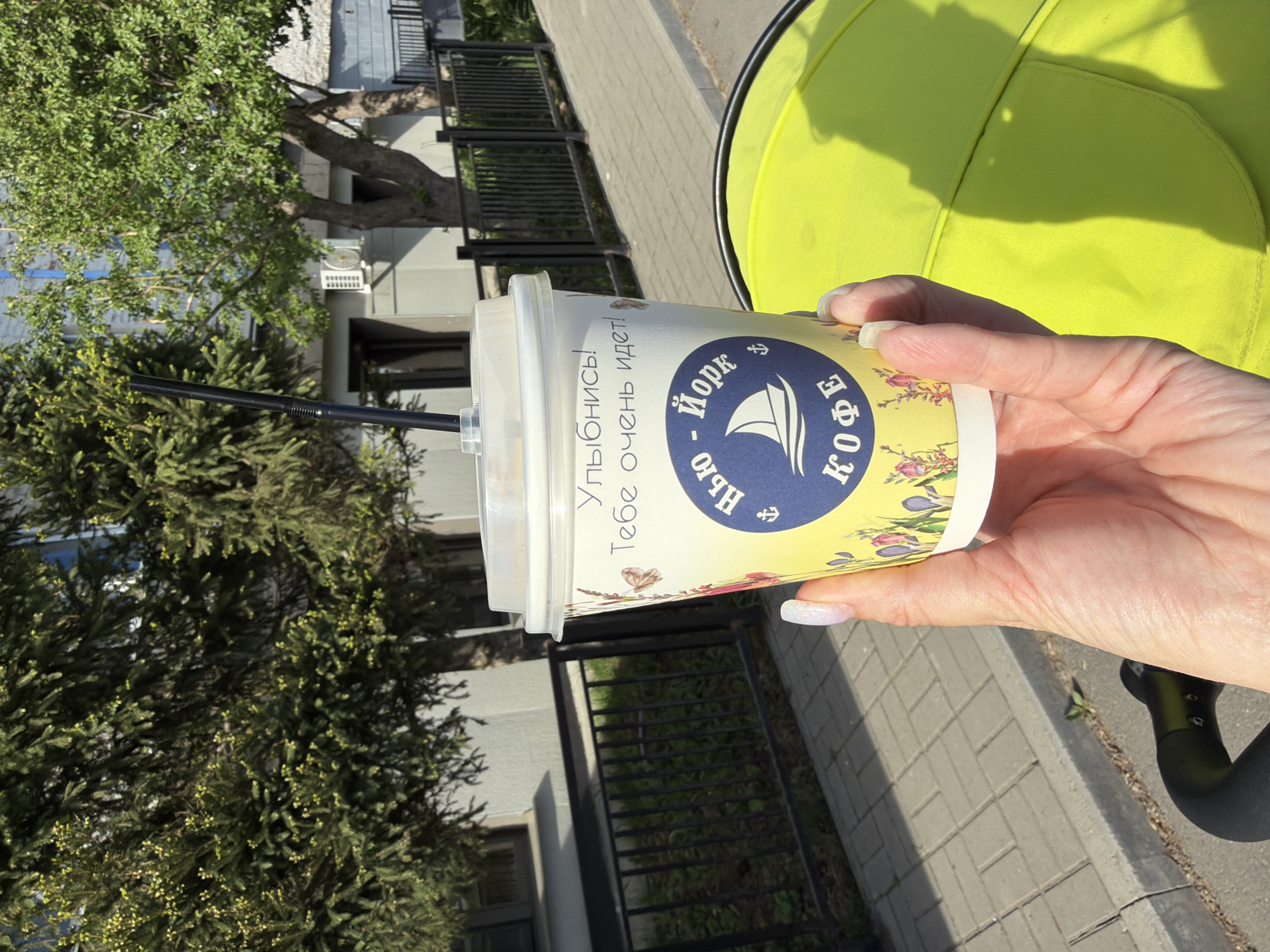 A person holds a cup of "York Coffee" in their hand, positioned near a stroller with a bright yellow interior. A black straw protrudes from the cup. The setting appears to be outdoors, likely in Votkinsk, Russia, as indicated by the coffee cup branding. The background includes a sidewalk paved with grey bricks, a green hedge, and a wrought iron fence. Parts of a building and trees are also visible.