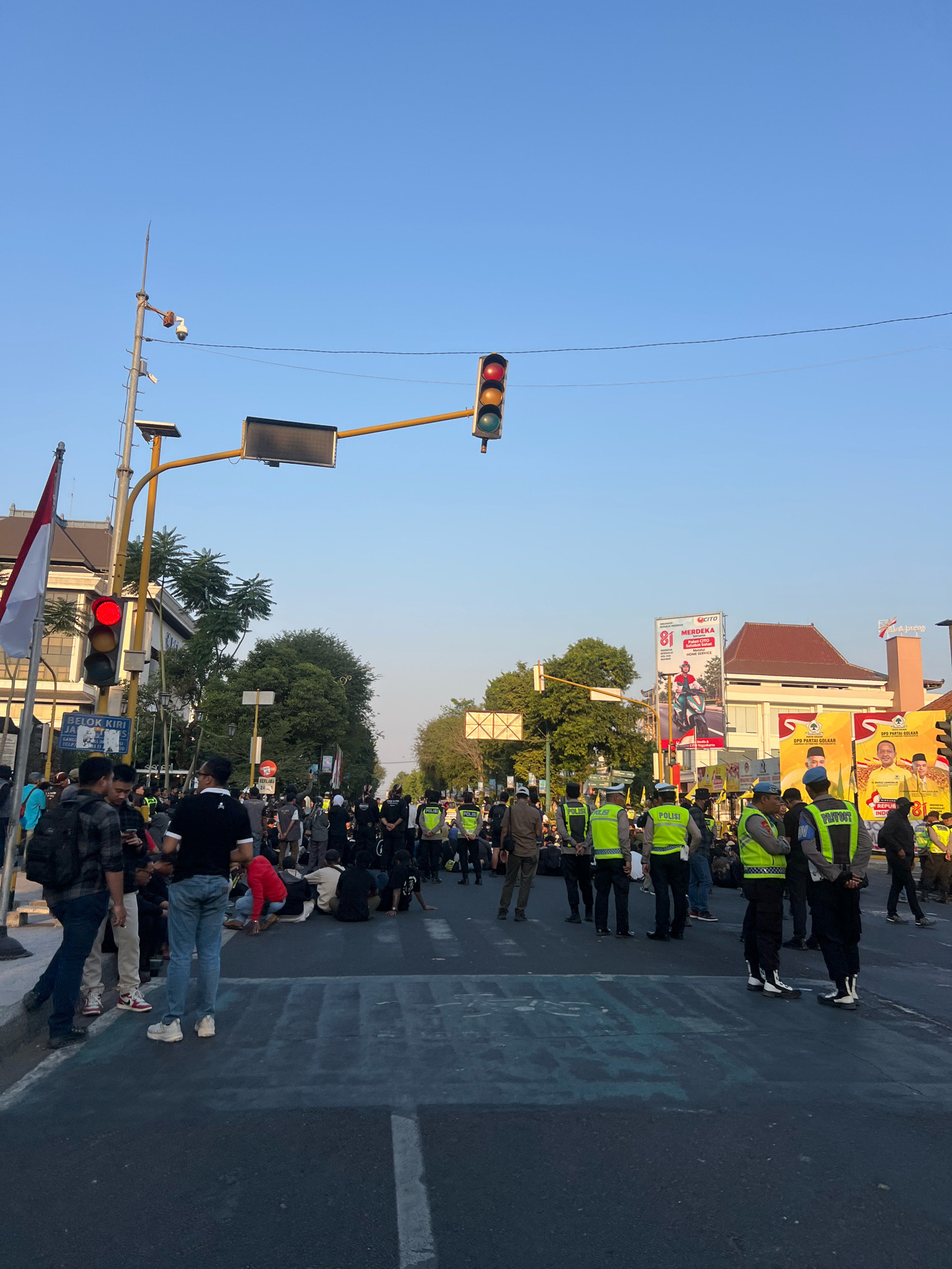 The image depicts a street scene in Yogyakarta, Indonesia, likely during the daytime given the clear blue sky and the presence of sunlight casting shadows. In the foreground, a street intersection is occupied by a group of people, some sitting on the asphalt and others standing. A significant number of individuals in high-visibility yellow vests with the word "POLISI" (Police) are present, indicating a law enforcement presence. Some of these police officers are wearing dark uniforms underneath their vests, and a few are wearing light blue helmets.
Traffic lights are visible overhead, with one red light illuminated, suggesting that traffic is currently halted. There are also traffic signs and what appears to be a large digital billboard or screen. Indonesian flags are visible, one prominently displayed on a flagpole on the left side of the image. Buildings line the street, including one with a reddish-brown tiled roof. Banners with campaign imagery, possibly for a political party, are displayed on the side of a building in the background. The street is paved with asphalt and has white lane markings. The overall atmosphere appears to be one of a demonstration or a controlled gathering, with police maintaining order.