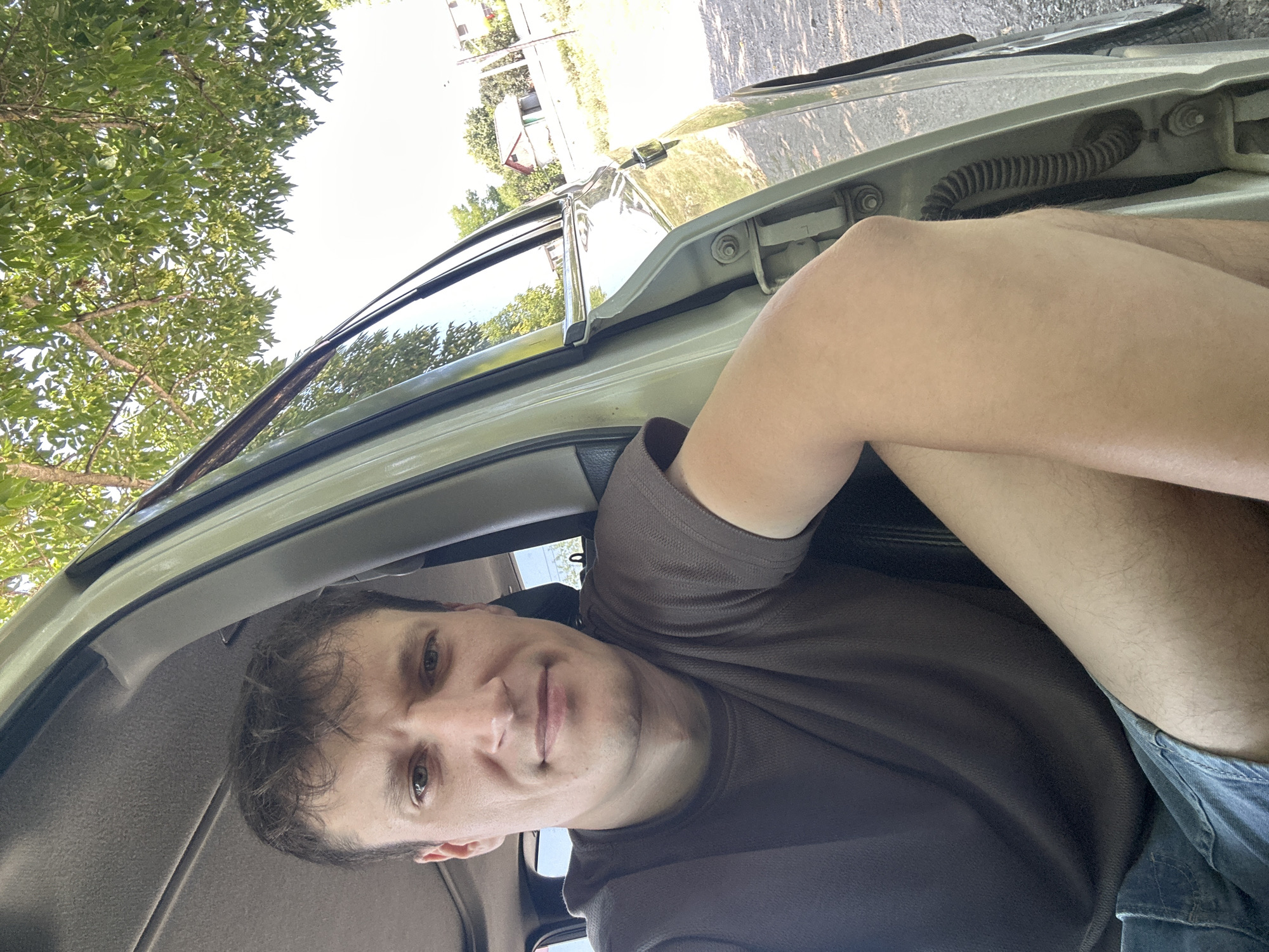 A man is sitting in the open driver's side door of a light-colored car, looking towards the camera. He has short, brown hair and is wearing a dark brown t-shirt and blue denim shorts. His left arm is resting on his leg. The car appears to be parked on a gravel surface next to a grassy area. In the background, there are trees with green leaves and a few houses with red roofs, consistent with a rural or suburban setting in Makiv, Ukraine. The lighting suggests it is daytime, likely a sunny day.