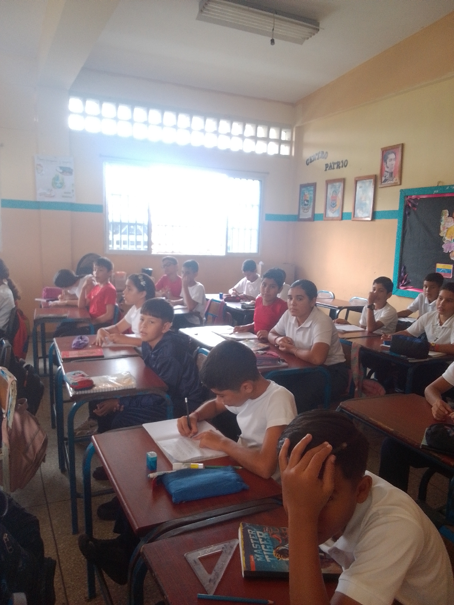 The image depicts a classroom scene in Santa Ana, Venezuela, likely during the daytime due to the bright natural light streaming through the windows. Several young students, appearing to be of primary or early secondary school age, are seated at individual desks. They are mostly engaged in activities related to their studies, with some looking towards the front of the classroom, others at their books or notebooks, and one student is seen writing.
The classroom appears functional, with rows of wooden desks and chairs. There is a large window at the back of the room, partially covered by blinds, letting in significant light. On the walls, there are several framed pictures or portraits, including what seems to be a national hero or historical figure. Text is visible on the wall above some of the framed pictures, reading "CENTRO PATRIO". There is also a bulletin board or display area on the far right wall with colorful decorations and possibly some student work or announcements.
The students are dressed in casual clothing, with many wearing white shirts. Their expressions and postures suggest a typical school day, with some appearing attentive and others perhaps a bit tired or contemplative. One student in the foreground is resting their head in their hand, possibly indicating fatigue or concentration. Objects on the desks include notebooks, pens, pencils, a ruler, and a book with the title "MASTER TIGER" visible. The overall atmosphere suggests a regular school day in session.