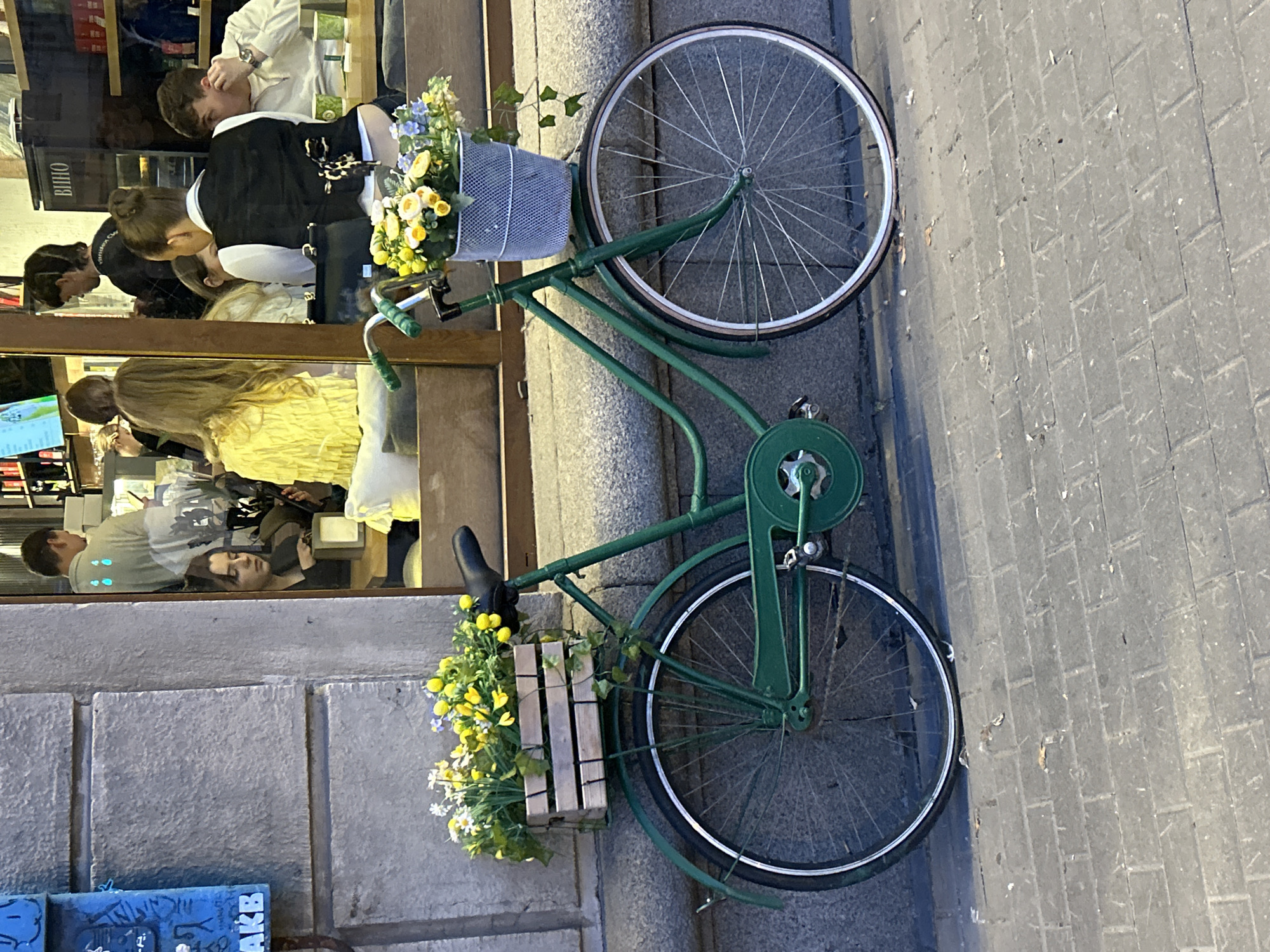 A green bicycle with a flower basket on the handlebars is parked on a cobblestone street in front of a building with a large window. The window reflects people inside a cafe or shop. The bicycle has a wooden crate of yellow and white flowers attached to the rear rack, and a white mesh basket of flowers on the handlebars. The building facade is made of stone blocks, and a blue mailbox is visible on the left side of the image. The scene is illuminated by natural light, suggesting it is daytime.