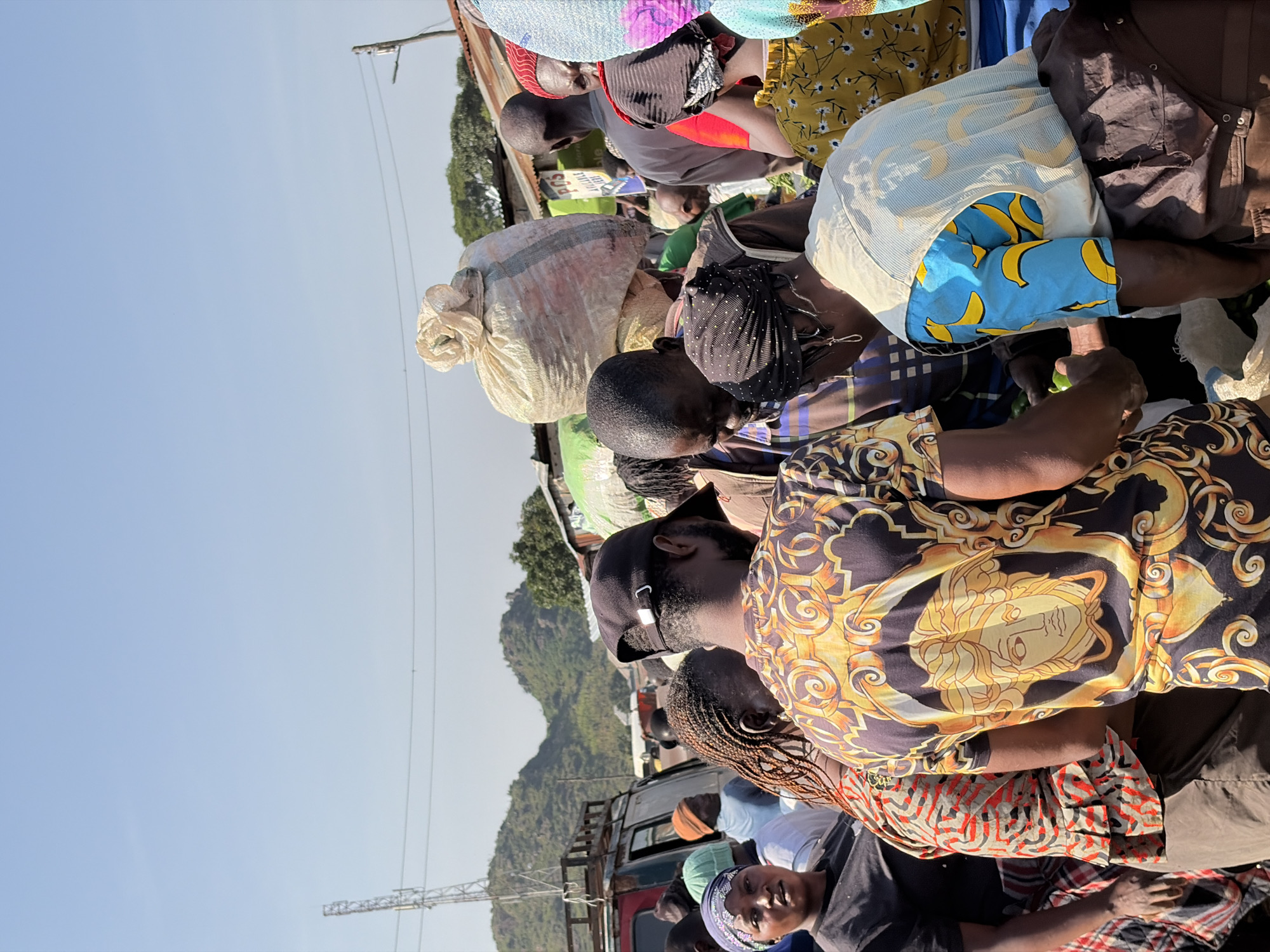 A medium shot captures a bustling outdoor market scene in Gura Top, Nigeria, under a clear blue sky. The foreground is dominated by a group of individuals, their faces mostly obscured as they engage in commerce. One person, seen from behind, wears a black baseball cap and a black shirt adorned with a gold Versace-like pattern featuring a Medusa head. To their left, a woman with braided hair wears a red and black patterned shirt. Further left, a woman in a blue headscarf and black t-shirt looks towards the camera. A large, bulging sack of what appears to be produce or grains is prominently displayed above the heads of the crowd. In the background, a red vehicle with a roof rack is parked, and beyond that, rolling green hills rise against the horizon, with a cellular tower visible on the left. The environment suggests a lively marketplace with an abundance of goods and a strong sense of community interaction.