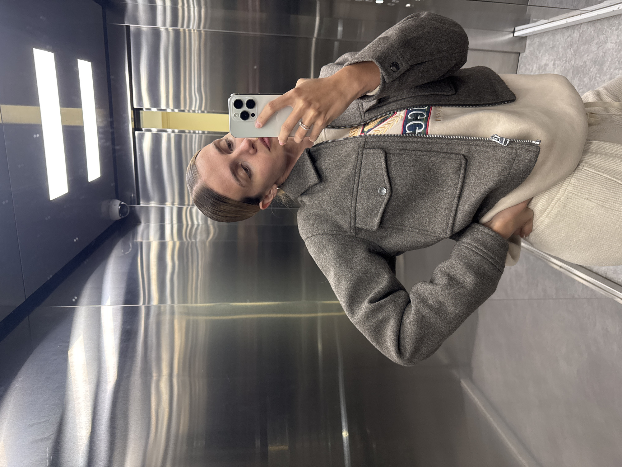 A person is taking a selfie in an elevator. The person is fair-skinned and wearing a gray jacket over a light-colored sweater. They are holding a white smartphone in their left hand, its camera facing them. The elevator has a shiny, metallic interior with vertical panels and a light fixture consisting of two elongated rectangular lights is visible on the left. The city and specific location are not discernible from the image.