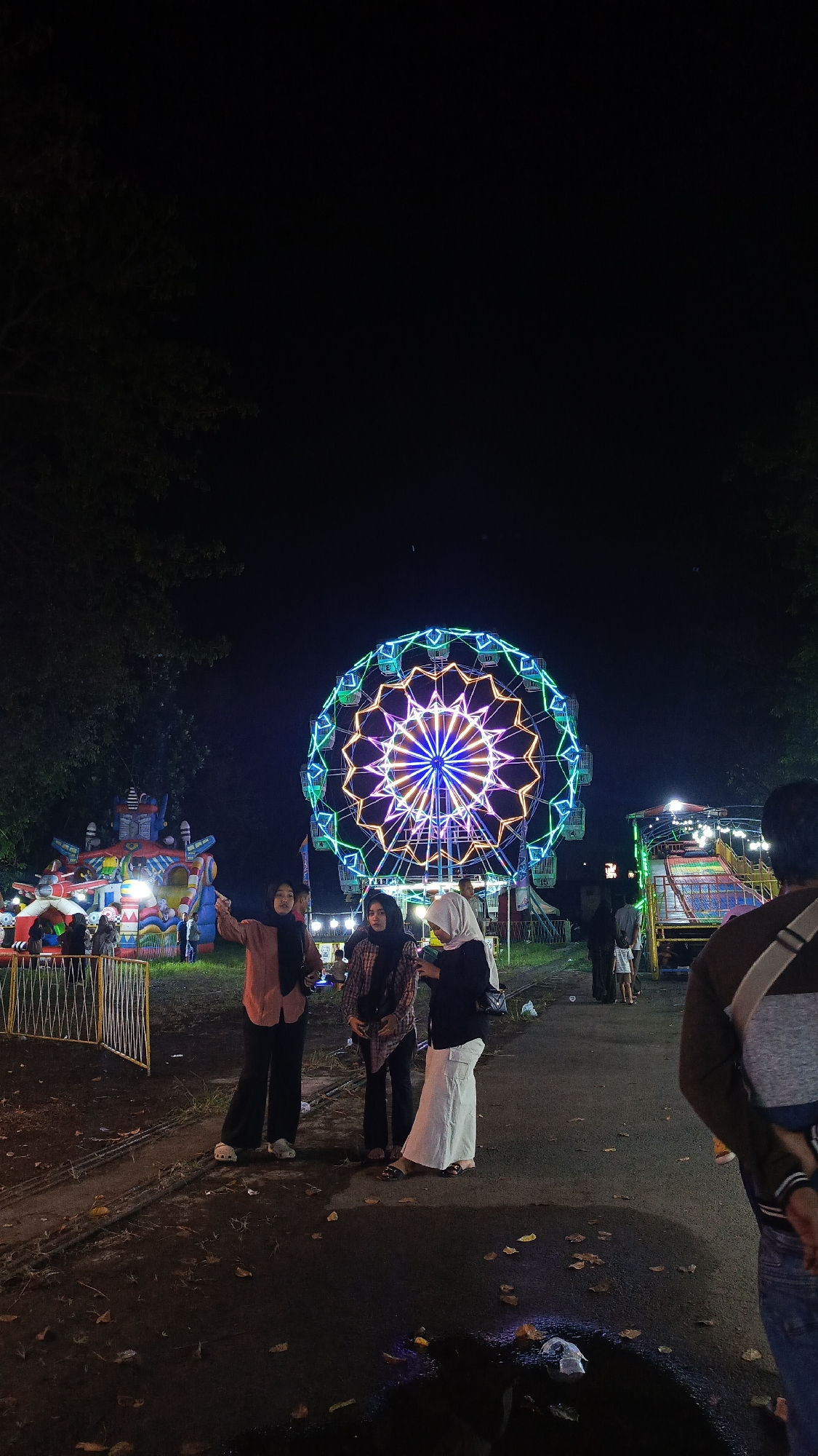 The image depicts an outdoor scene at night in Tulangan, Indonesia, featuring a brightly lit Ferris wheel as a central element. In the foreground, three women are standing on a paved surface. The woman on the left is wearing a pink top with a black hijab, dark pants, and white shoes, with her right arm raised. The woman in the middle is wearing a plaid top with a black hijab and dark pants. The woman on the right is wearing a white hijab with a black top and white skirt.
Behind the women is the Ferris wheel, illuminated with green, blue, and purple lights. The ground is dark and appears slightly wet, with fallen leaves scattered on the pavement.
Other carnival rides and attractions are visible in the background. The lighting suggests a festive atmosphere, likely an amusement park or carnival setting. There are other people visible in the background, adding to the scene's activity.