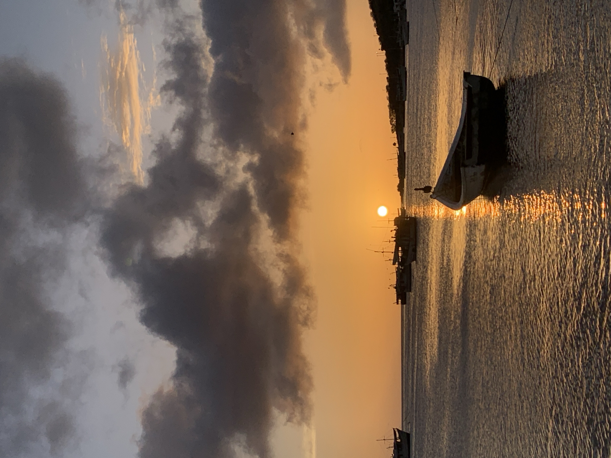The image depicts a tranquil scene at sunset in Juan Griego, Venezuela. The sky is ablaze with a warm orange and yellow hue, transitioning to softer blues and grays as dramatic, dark clouds drift across the horizon. The sun, a bright disc, is low in the sky, casting a shimmering golden reflection on the calm water.
Several boats are anchored in the bay. In the foreground, a small, weathered rowboat rests gently on the water, its dark hull contrasting with the bright reflections. A pelican is perched on the edge of this rowboat, its silhouette distinct against the sunset. Further out, larger fishing vessels are scattered across the water, their masts and structures silhouetted against the vibrant sky. The shoreline in the distance is lined with lush greenery and hints of buildings, suggesting the presence of the town of Juan Griego. The overall atmosphere is one of peaceful serenity as day transitions into night.