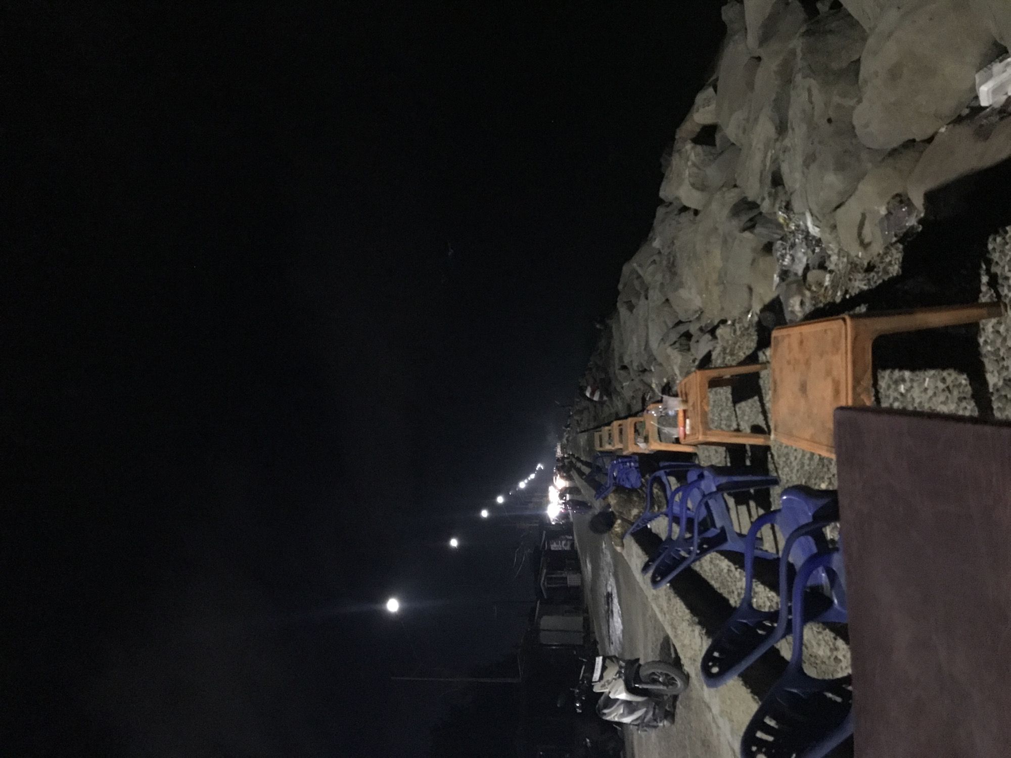 The photograph captures a night scene at a seaside location in Lhokseumawe, Indonesia. The camera is oriented alongside a promenade next to a shoreline composed of large rocks and gravel. The scene is illuminated by a series of streetlights, creating bright points of light along the walkway.
The immediate foreground of the photograph shows a row of plastic blue chairs and small brown tables, presumably for outdoor seating at a seaside establishment. A few motorbikes are parked along the edge of the promenade. A person is partially visible amidst the chairs, possibly sitting or working in the area.