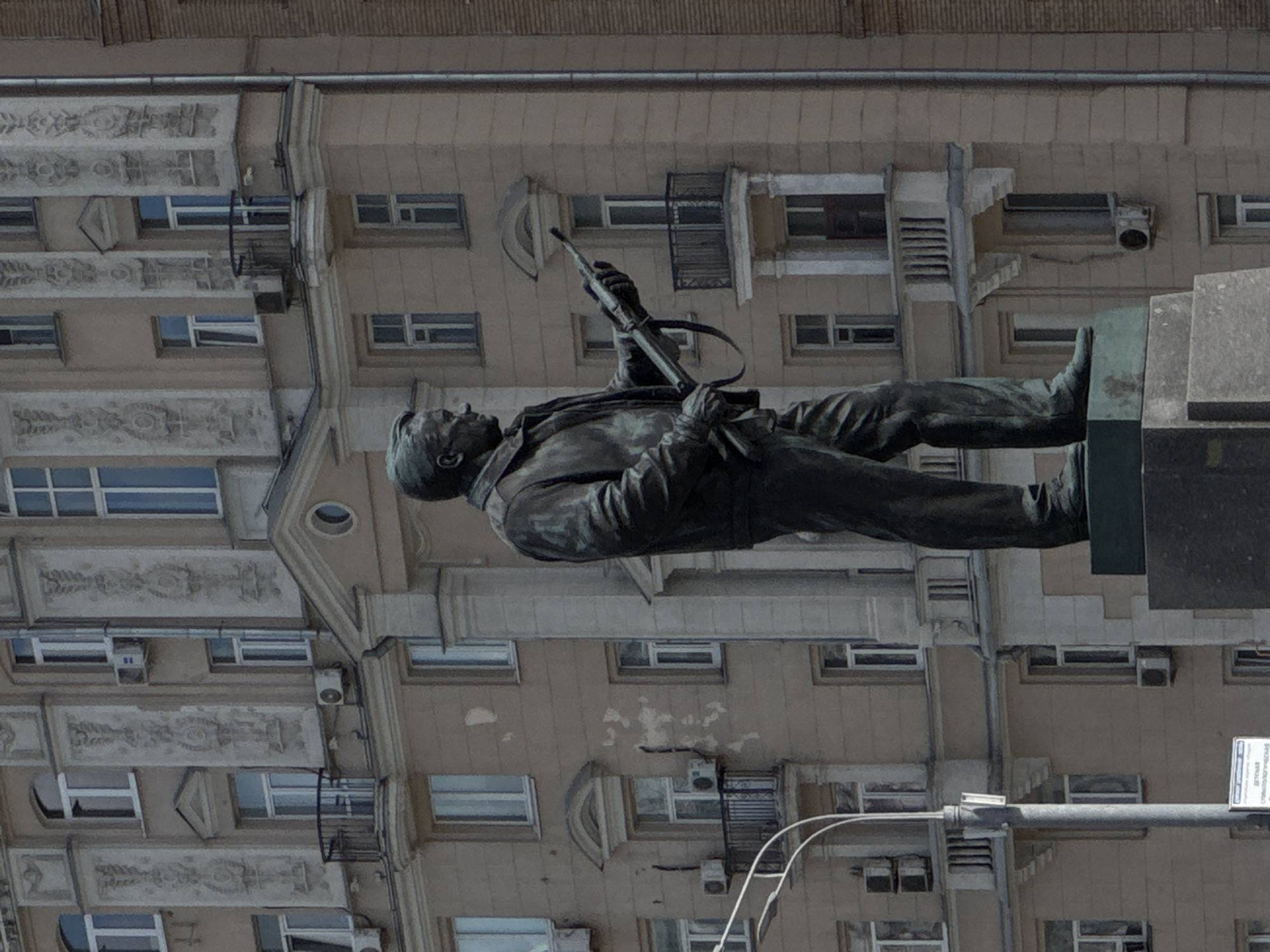 The image shows a bronze statue of a man holding a rifle, standing in front of a large, ornate, light-brown building with numerous windows and balconies. The statue is positioned to the right of the frame, facing left. The building's architecture suggests it is a pre-war Stalinist era building, typical of Moscow, Russia. The statue itself appears to be a monument. The building features decorative elements around the windows and between floors. A street lamp is visible on the left side of the image. The statue's pose and the rifle suggest it might commemorate a soldier or a historical figure. The lighting indicates it is daytime.