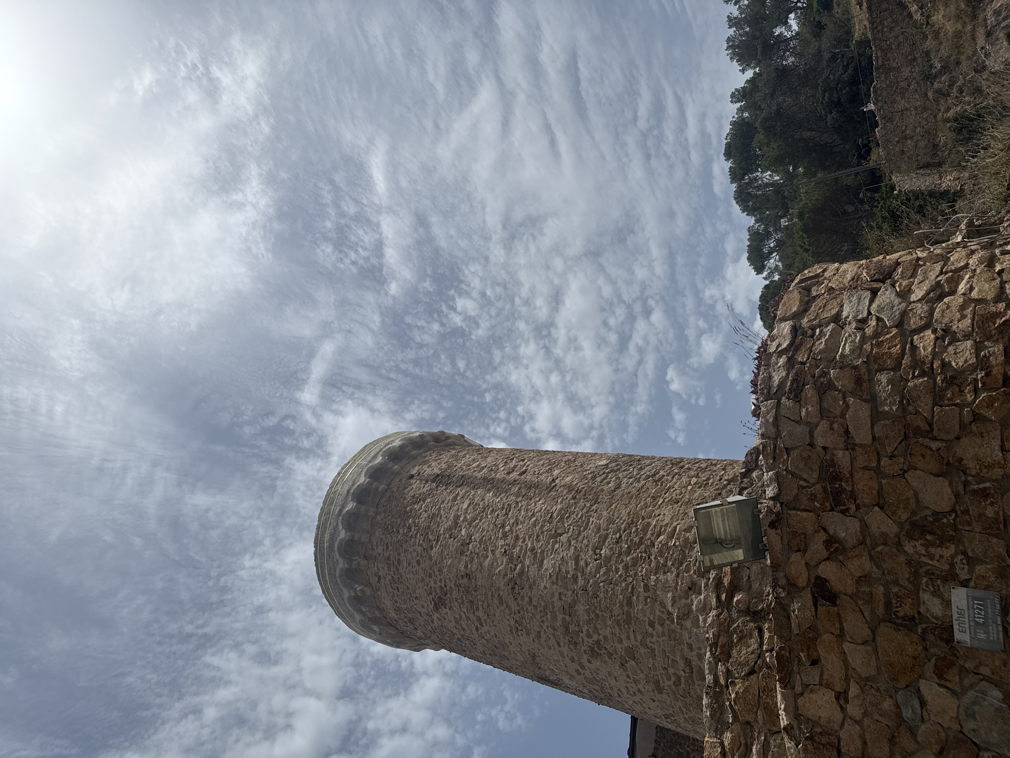 This is a low-angle, full-frame shot of the Torre dels Moros, a medieval watchtower located in Tossa de Mar, Spain. The tower is constructed of rough-hewn stone blocks and has a cylindrical shape, tapering slightly towards the top. It is capped with a decorative cornice and a protective netting. The tower is situated on a stone-paved terrace, with a metal floodlight attached to the wall in the foreground. Behind the tower, a dense forest of green trees covers a hillside, with a glimpse of a clear blue sky with wispy clouds overhead. A sign with "enher N° 41271" is visible on the stone wall near the bottom left of the frame.