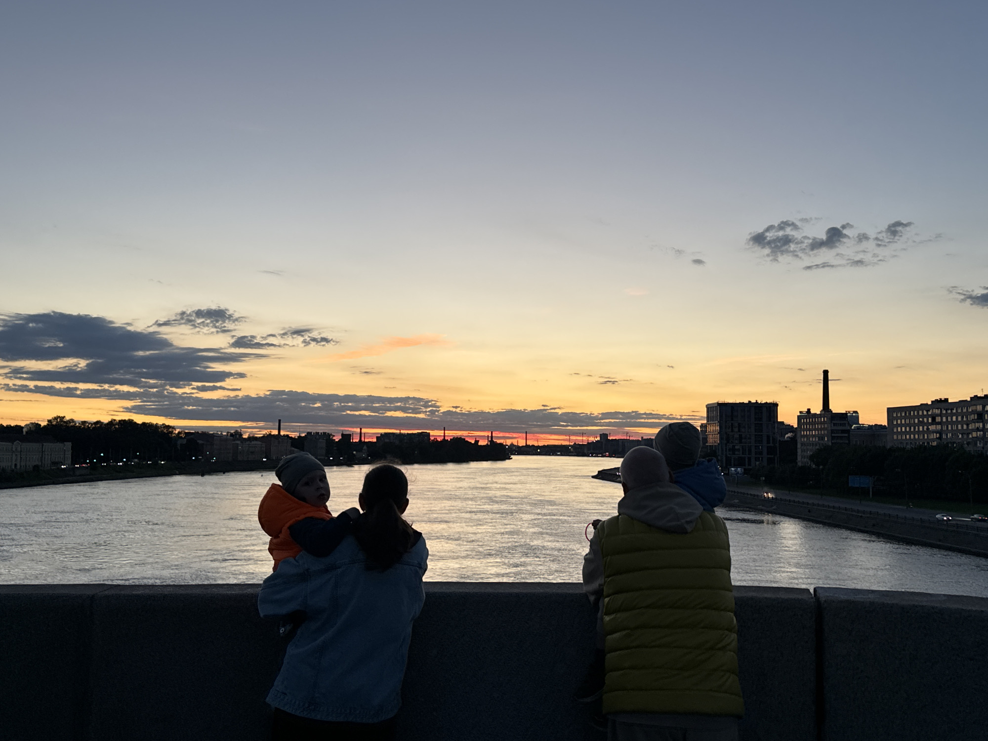 This image depicts a family enjoying a sunset over a river in Staraya, Russia. In the foreground, silhouetted figures of people are visible from behind, facing the water and the cityscape. A woman is holding a baby on her left shoulder, and another person stands to her right, also facing the sunset. The baby is wearing an orange jacket and a gray hat. The woman is wearing a blue denim jacket. The person to her right is wearing a yellow puffer vest over a gray hooded sweatshirt. The river occupies the middle ground, reflecting the warm colors of the sky. The background shows the silhouette of buildings and trees lining the riverbank, with a tall smokestack visible on the right side of the frame. The sky is a gradient of soft blues and oranges, with some clouds scattered across it, indicating the end of the day. The overall impression is one of a serene family moment against a beautiful natural backdrop in a Russian city.