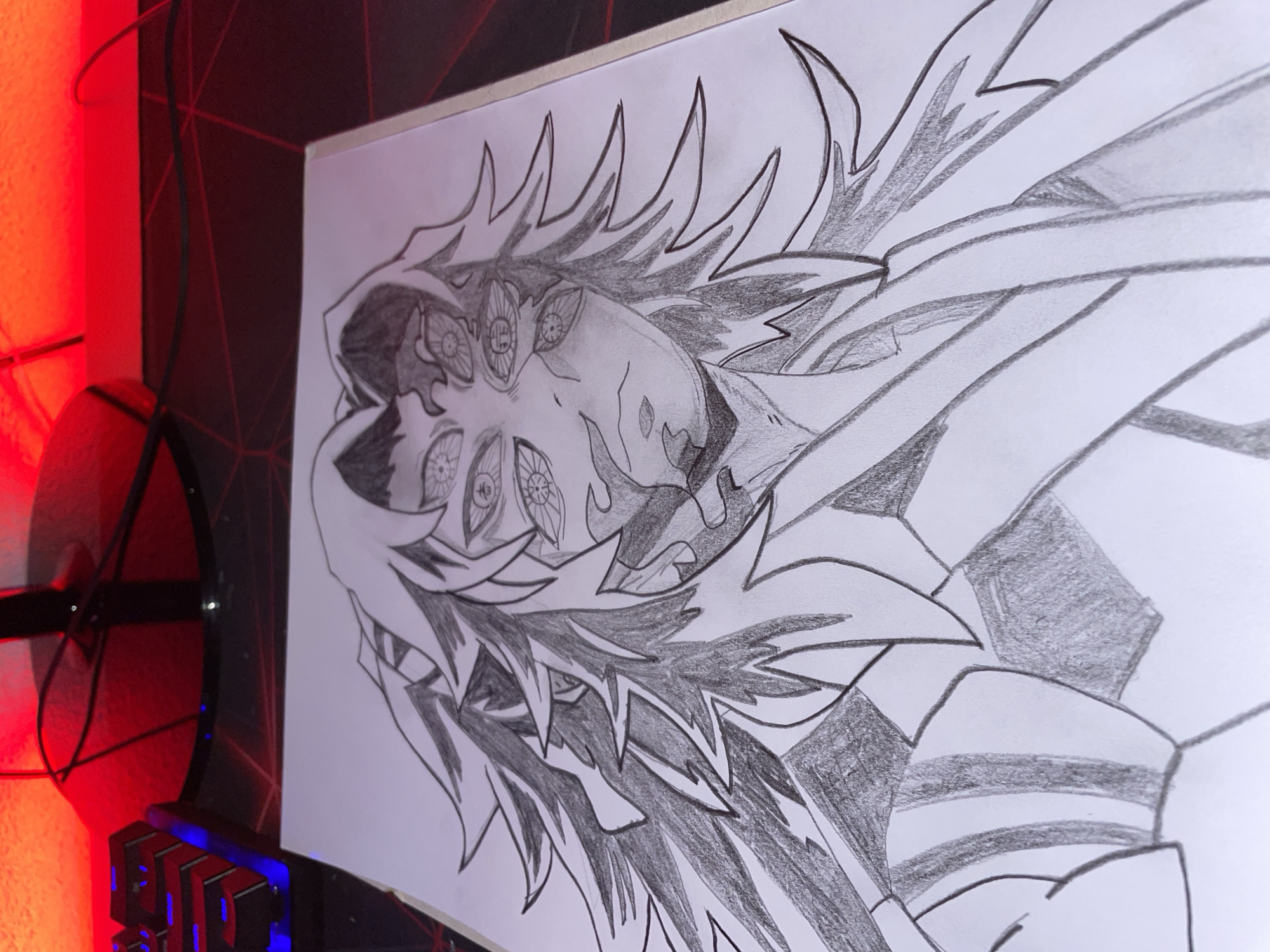 The image shows a pencil drawing of a character with multiple eyes and spiky hair. The drawing is on a white sheet of paper. In the background, there is a black computer mouse pad with a keyboard partially visible in the lower left corner. There is also a fan and a red light source.