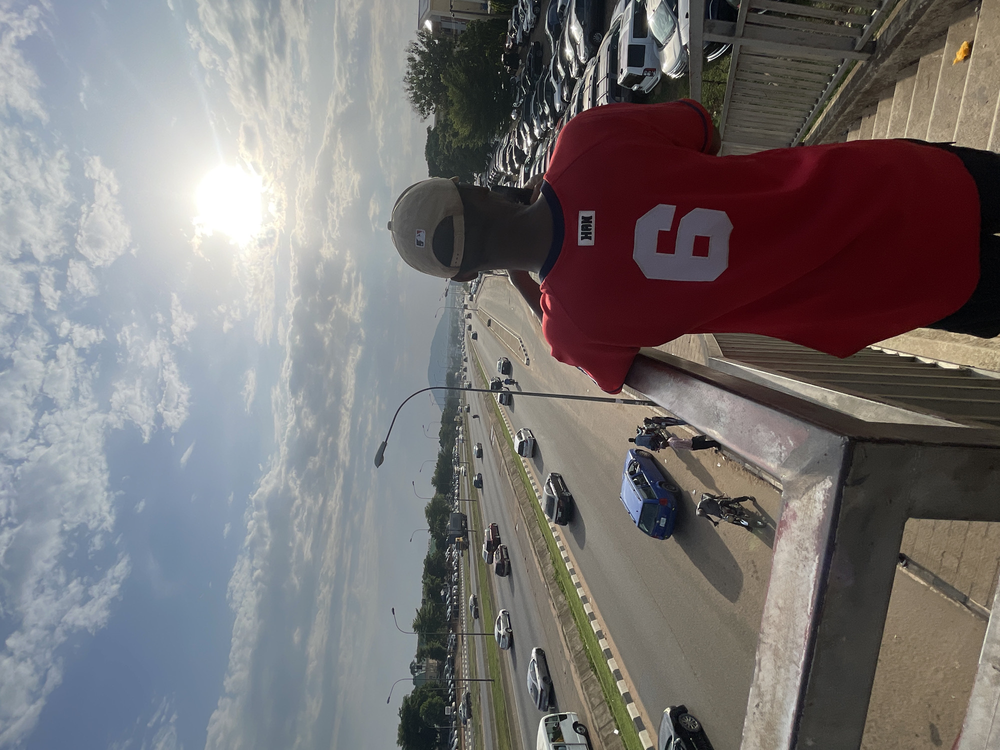 A person in a red jersey and tan baseball cap stands on a pedestrian overpass in Gwarinpa, Nigeria. The person looks out over a busy highway with cars and motorcycles. A blue car and several motorcycles appear to be involved in an accident on the highway. Parked cars line the side of the highway. The sky is bright with clouds.