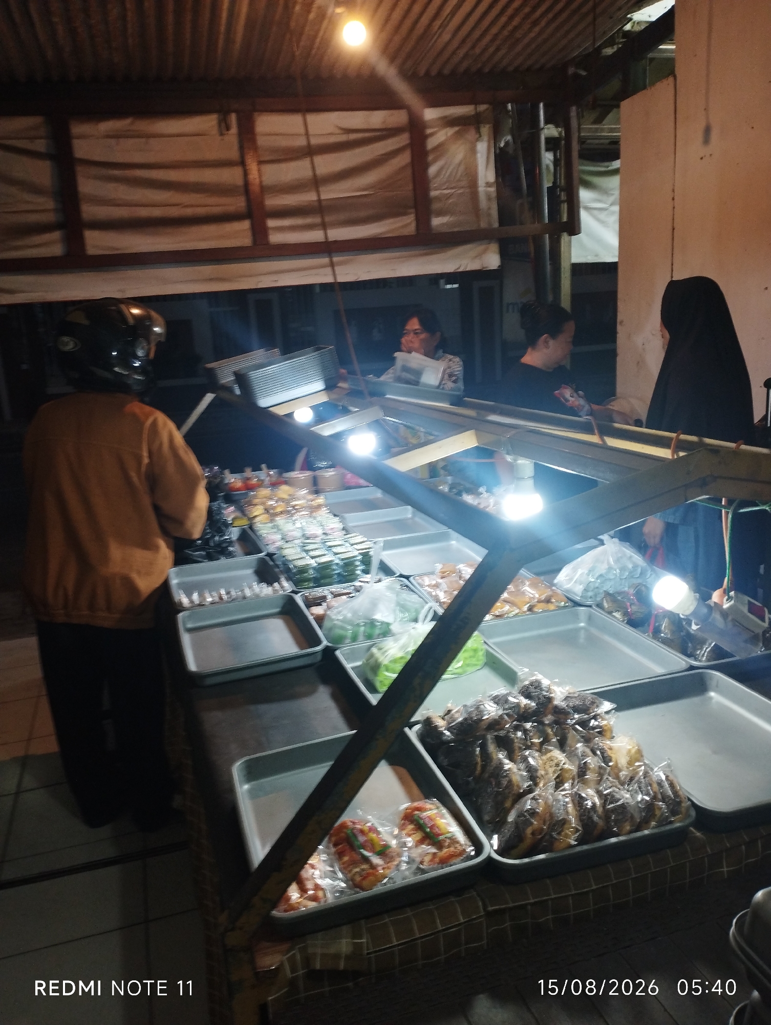 This image depicts a food stall at night, likely in Karawang, Indonesia, as indicated by the timestamp. The stall appears to be outdoors, with a corrugated metal roof and overhead lighting. The foreground features several metal trays filled with various food items, including what appear to be wrapped snacks or pastries, some loose food items in plastic bags, and possibly some fresh produce. A person wearing a helmet and an orange jacket is standing on the left, facing the stall and seemingly engaged in purchasing food. To the right, two women are visible. One, wearing a black hijab, is standing behind the stall, possibly attending to customers, while the other, in a black top, is looking at something on the stall. The overall impression is a bustling night market scene.