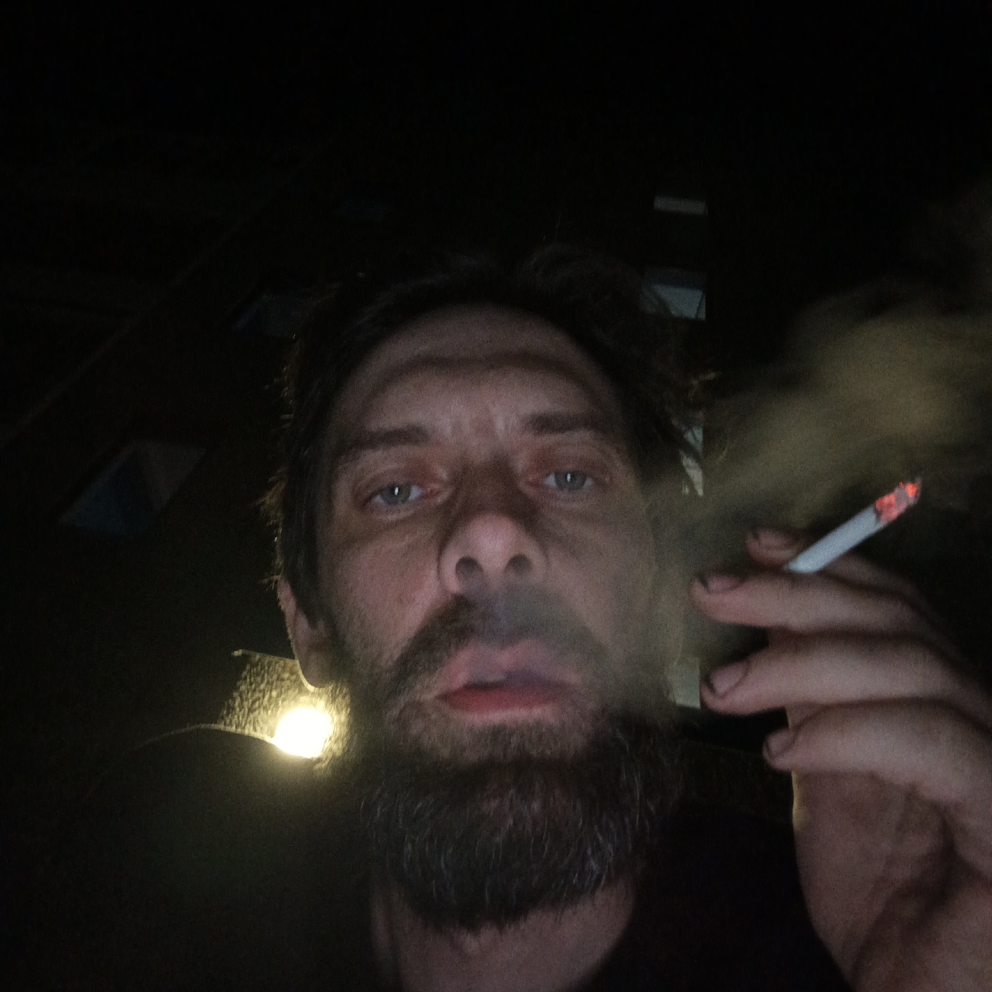 A low-angle, dimly lit close-up selfie of a Caucasian man with fair skin, light blue eyes, dark brown hair, and a full, unkempt beard. He is holding a lit cigarette in his right hand, from which smoke is emanating. The man is looking directly at the camera. The background is dark, with vague, indistinct shapes suggesting buildings and a light source partially visible in the lower left. The location, Марковский, Russia, is not discernible from the visual information within the image.