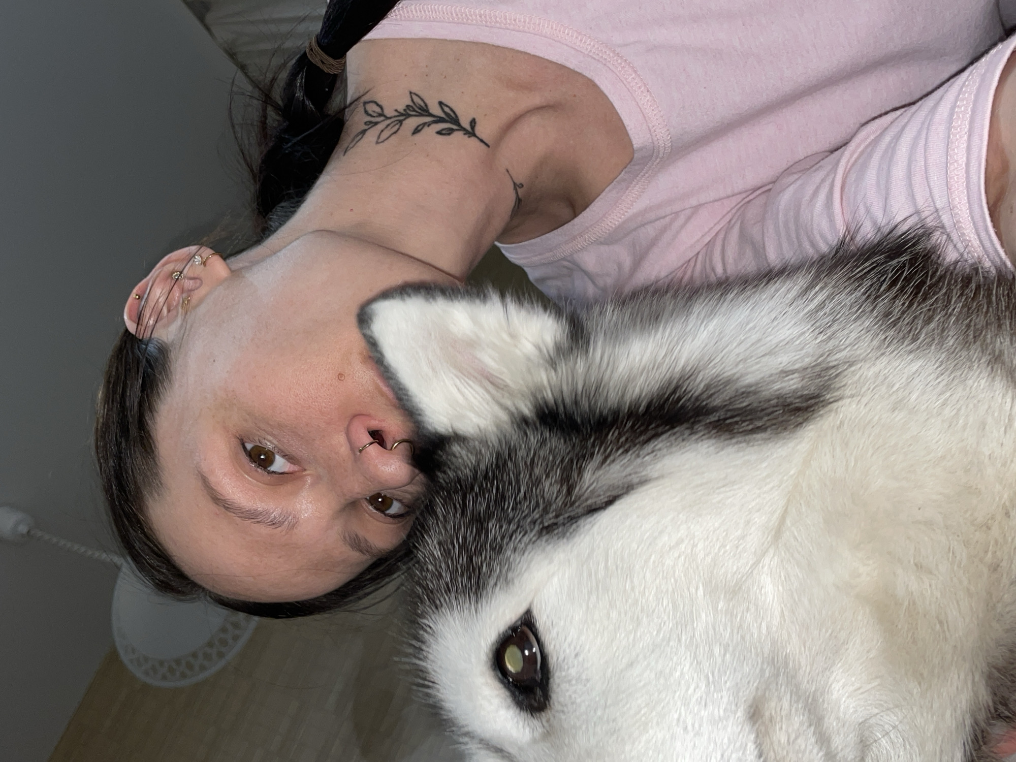 A woman with dark hair, a nose ring, and multiple ear piercings is posing for a selfie with her white and grey Husky dog. She is wearing a light pink t-shirt and has a leafy branch tattoo on her collarbone. The setting appears to be indoors, based on the lighting and partial view of a light fixture on the ceiling. The location is unknown and there is no information in the image to suggest it is Izhevsk, Russia.