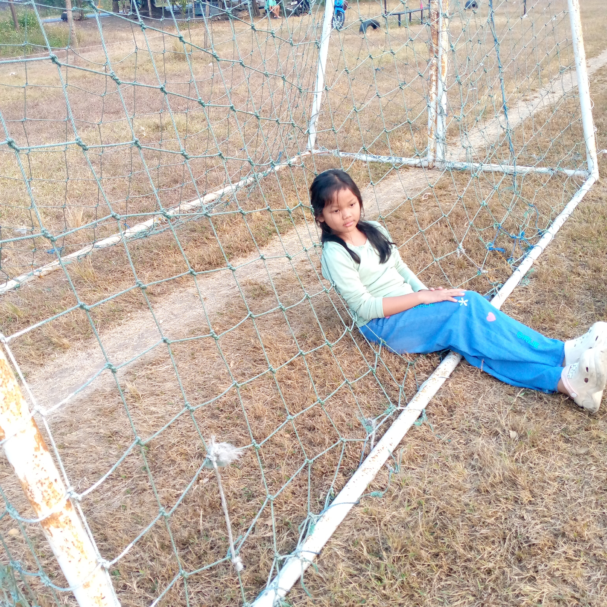 A young girl with dark hair is sitting on the ground, leaning against the netting of a soccer goal. She is wearing a light green long-sleeved shirt and blue pants. Her legs are extended in front of her. The ground is covered in dry grass, and a dirt path runs behind the goal. In the background, there are trees and some scattered objects, including what appears to be a bicycle and some benches. The scene is outdoors, likely on a sports field. The image is taken from a slightly elevated angle, looking down at the girl and the goal. The image is not taken in Pacing, Indonesia.