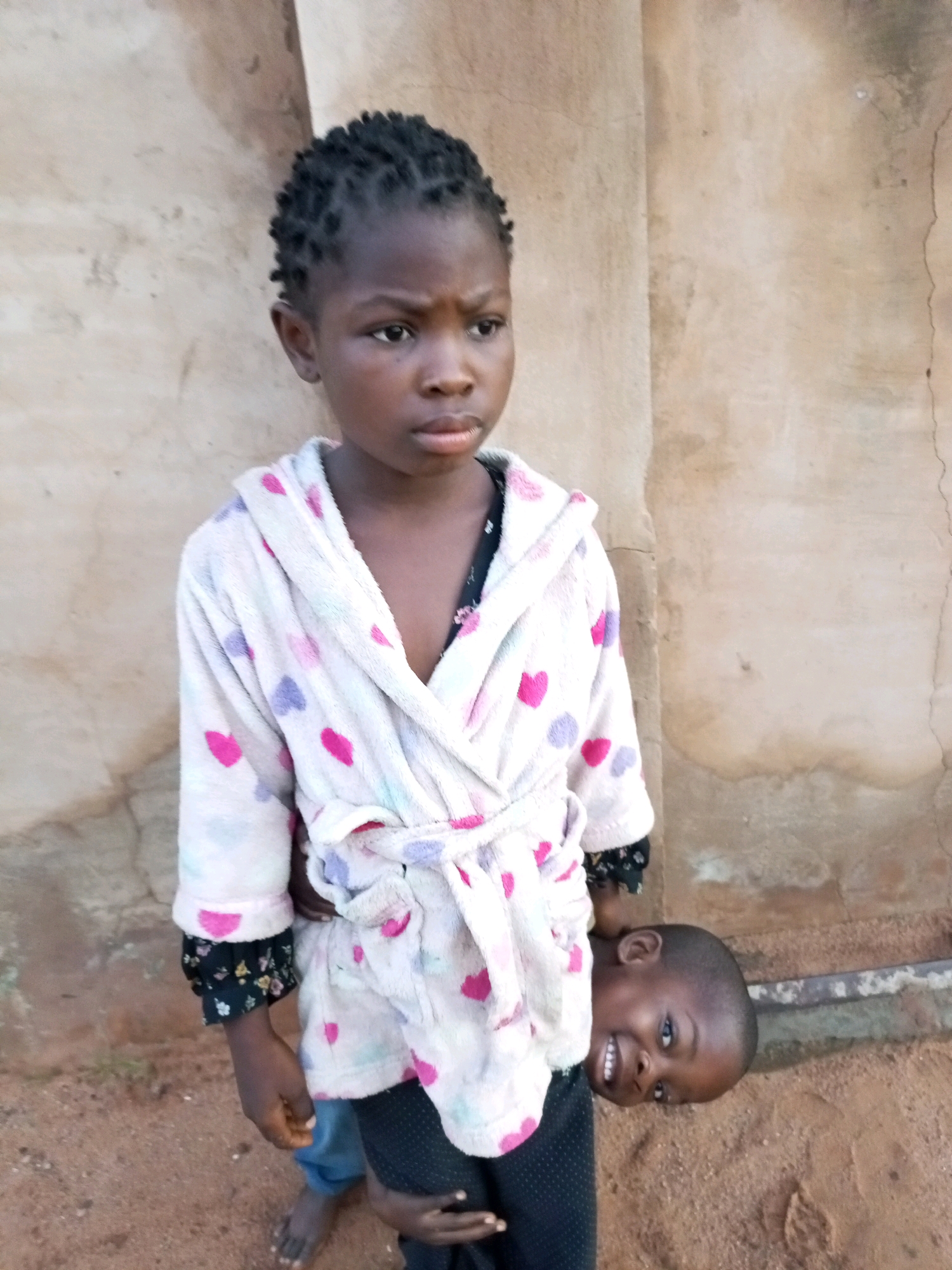 In Tempure, Nigeria, two young children stand against a textured, light brown wall. The child in the foreground, likely a girl, is facing slightly to the right, wearing a white bathrobe adorned with pink and purple hearts. Her hair is styled in small, dark braids. She is holding a younger child, possibly a boy, who is peeking out from under her arm, his head tilted and face turned towards the camera with a wide, toothy smile. The younger child is wearing dark clothing. The ground is covered with reddish-brown dirt. A thin, horizontal metal pipe runs across the background near the ground.