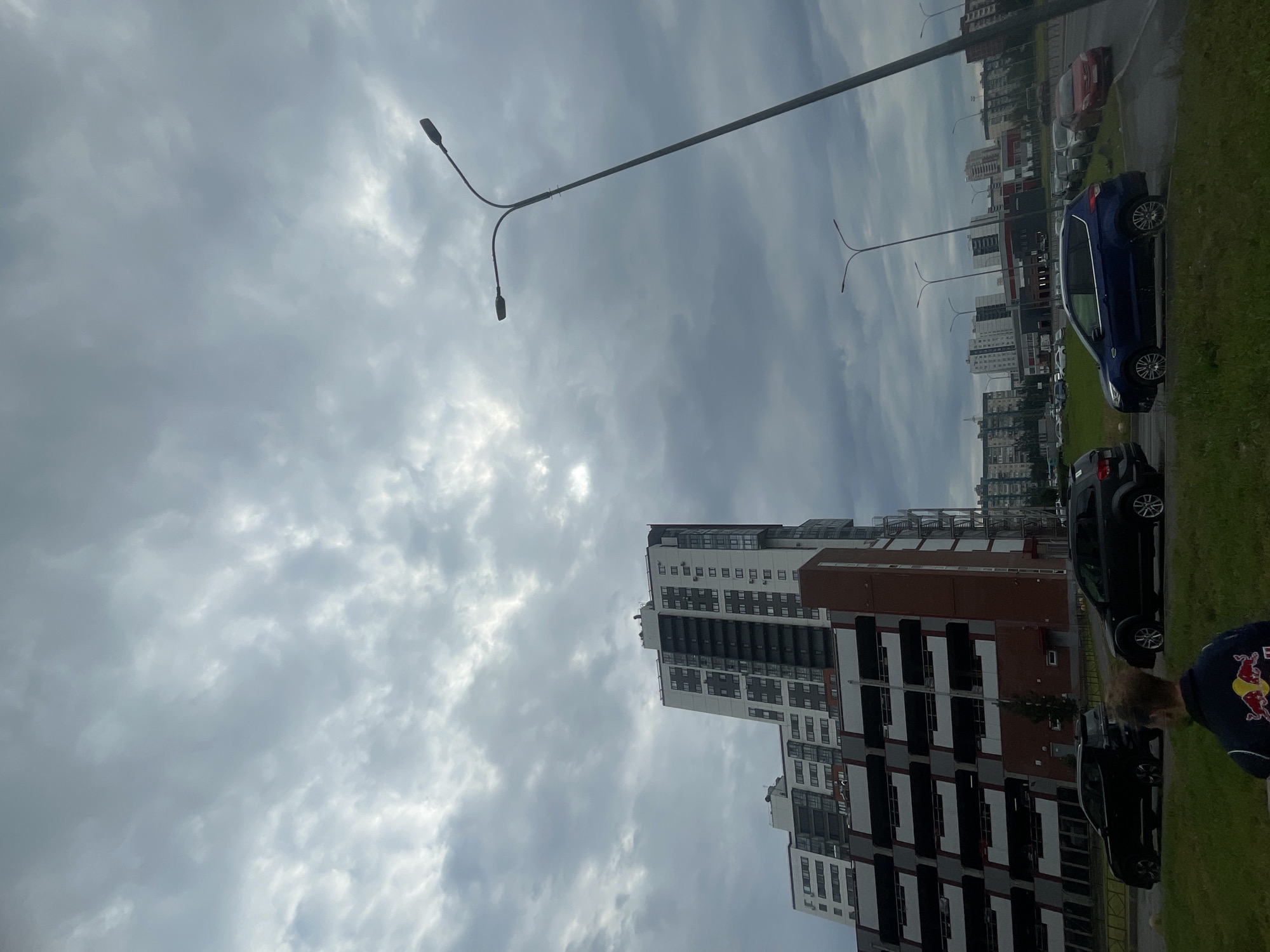 The image captures a cloudy, overcast day in an urban environment. In the foreground, a person is partially visible from behind, wearing a dark jacket with a red and yellow logo. Adjacent to the person, on a paved surface, are several parked vehicles, including a black SUV on the left and a black SUV in the middle. Further down the road, a blue SUV is visible, followed by other cars. In the background, a large multi-story building complex with a red brick and dark gray facade dominates the scene. This structure appears to be a parking garage or a mixed-use residential building. Beyond it, other residential buildings and some commercial structures are scattered across the landscape. Two tall streetlights with multiple lamps are prominent in the frame, one extending from the right side of the image towards the top, and another shorter one visible in the distance. The sky is filled with dense, gray clouds, with some lighter patches suggesting a break in the overcast conditions. The overall impression is of a typical suburban or city scene under a somber sky.