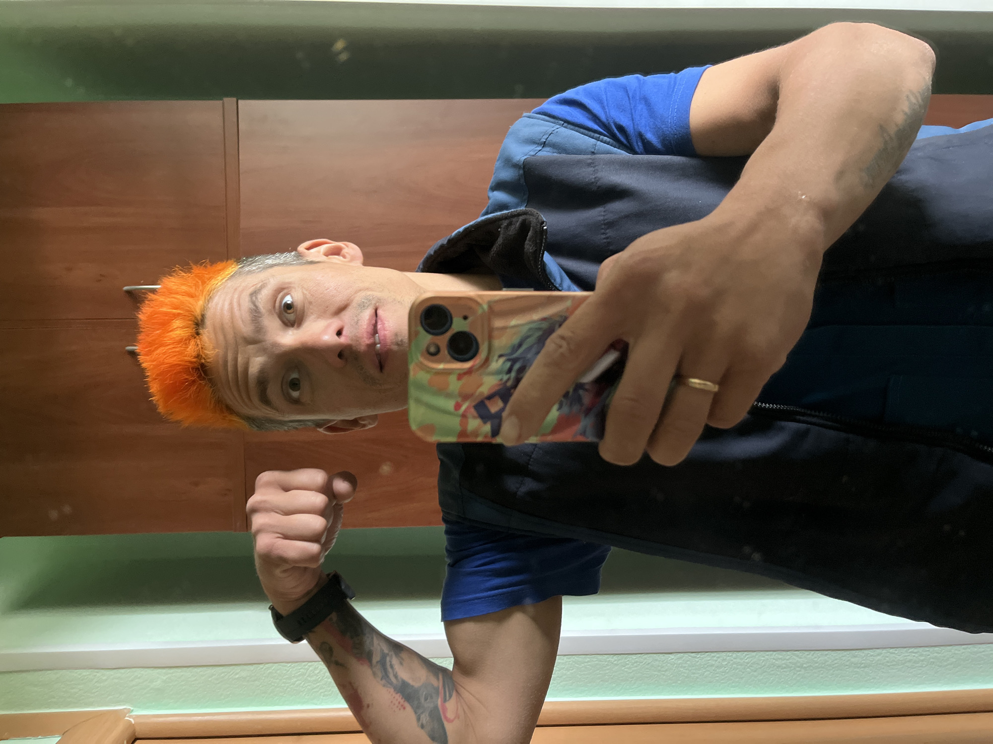 A dynamic mirror selfie taken indoors, rotated 90 degrees clockwise, captures a man with distinctive features. The subject, in what is likely Chaikovsky, Russia, has striking spiky hair that is bright orange on top and gray on the sides. He has light-colored, wide-set eyes, and a somewhat surprised or playful expression, looking directly into the smartphone's camera.
He is dressed in a royal blue t-shirt underneath a dark blue or black sleeveless vest or jacket. His left arm, visible in the foreground, is muscular and adorned with several tattoos, with his hand clenched into a fist. A dark digital watch is worn on his left wrist. His right hand holds a light-colored smartphone (an iPhone with a triple-camera system and a colorful, artistic case), which he is using to capture the image. A plain gold band ring is visible on the ring finger of his right hand.
The background indicates an indoor setting. Above the man's head (as oriented in the rotated image) are sections of what appear to be wooden cabinets or furniture with a smooth, reflective surface. Below him, a light green wall is visible, with a prominent white horizontal trim or ledge separating the upper wall from a darker area further down. The lighting suggests an interior, possibly artificial, environment. No specific time of day, weather, or visible text is discernible from the image.