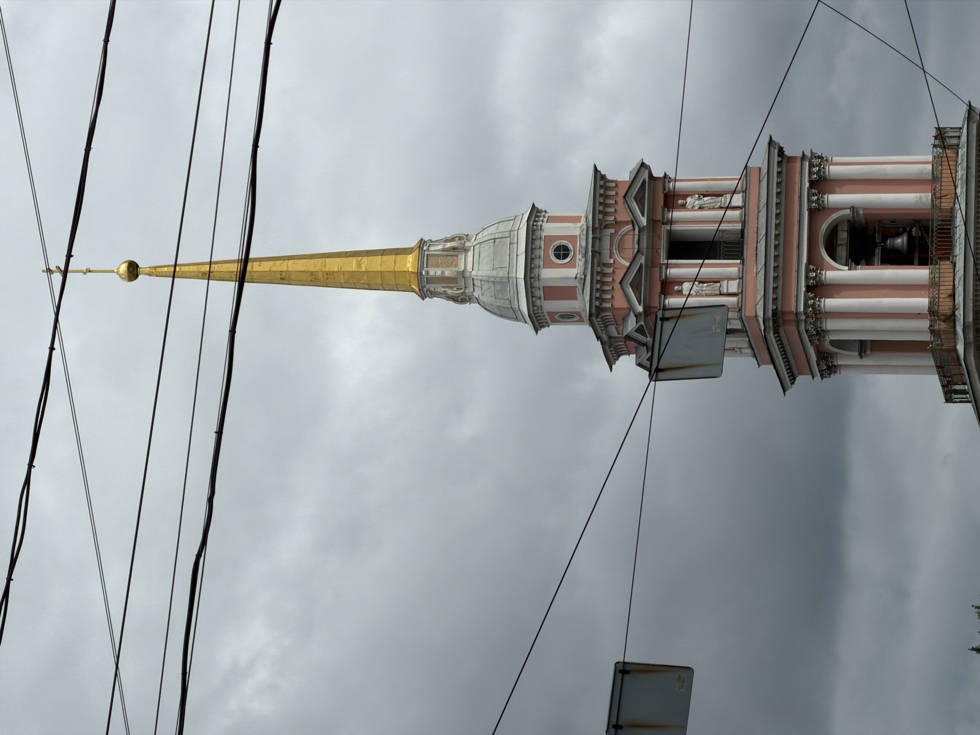 The image shows the bell tower of the Peter and Paul Cathedral in Saint Petersburg, Russia, under a cloudy sky. The tower is predominantly pink and white, with a tall, golden spire topped with a cross. Several power lines crisscross the frame, obscuring parts of the sky and the tower. The bell tower has architectural details such as columns, arched openings, statues, and a balcony. A large bell is visible within one of the arched openings. Two square signs, one on the left and one on the right, are also present, partially obscuring the view.