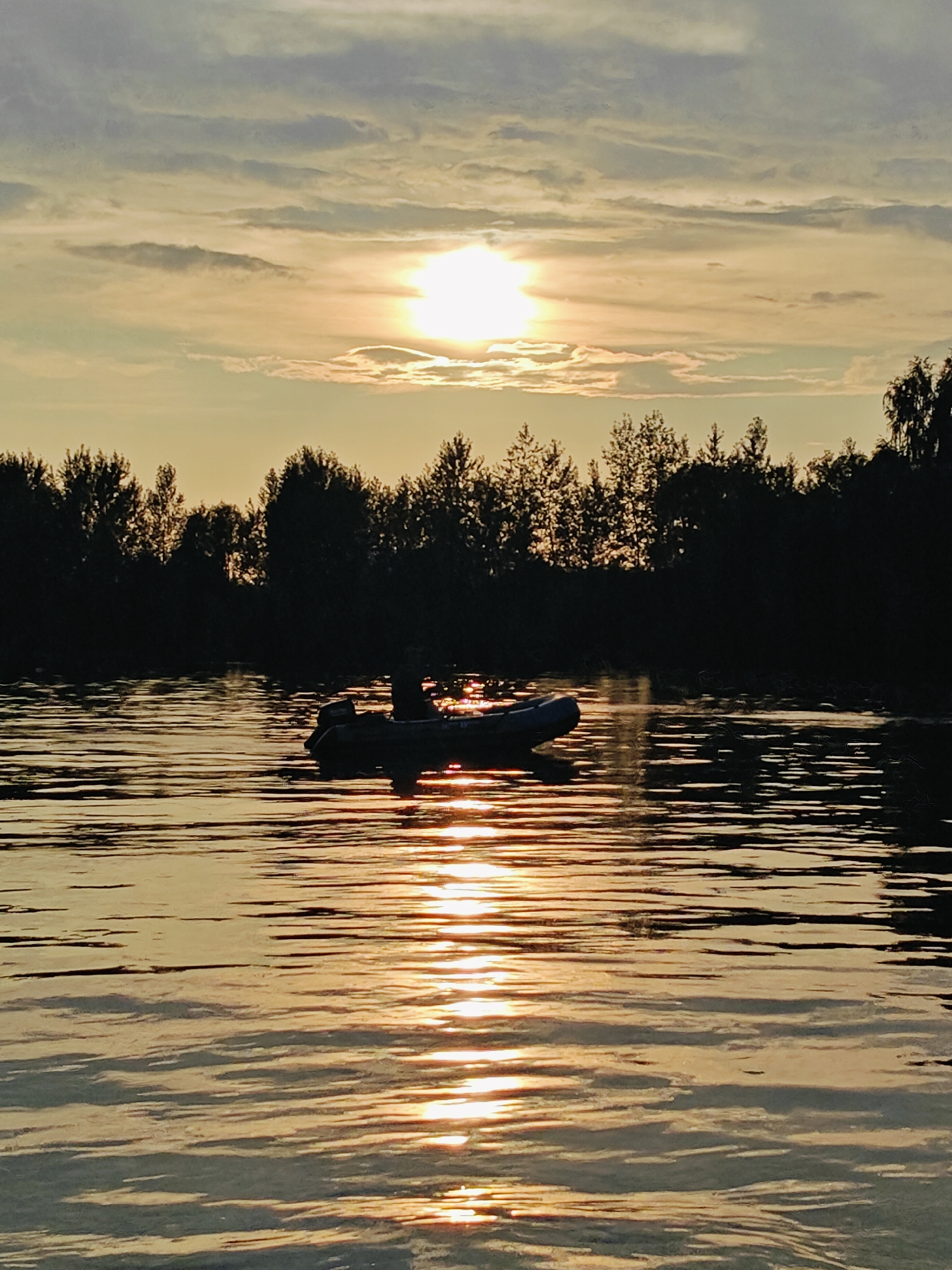 An outdoor, eye-level, medium shot captures a serene sunset over a body of water, possibly a river or lake, in Ol'khovskoye, Russia. The sky is painted with warm hues of yellow and orange, with the sun appearing as a bright orb partially obscured by wispy clouds. Silhouetted against the radiant sky is a dark treeline bordering the opposite bank, creating a stark contrast. In the foreground, the water reflects the vibrant colors of the sunset, creating a shimmering, golden surface. A small, inflatable boat with an outboard motor is visible in the middle ground, with a single person sitting in it, casting a faint shadow on the water. The overall atmosphere is peaceful and tranquil, evoking a sense of quietude and natural beauty.