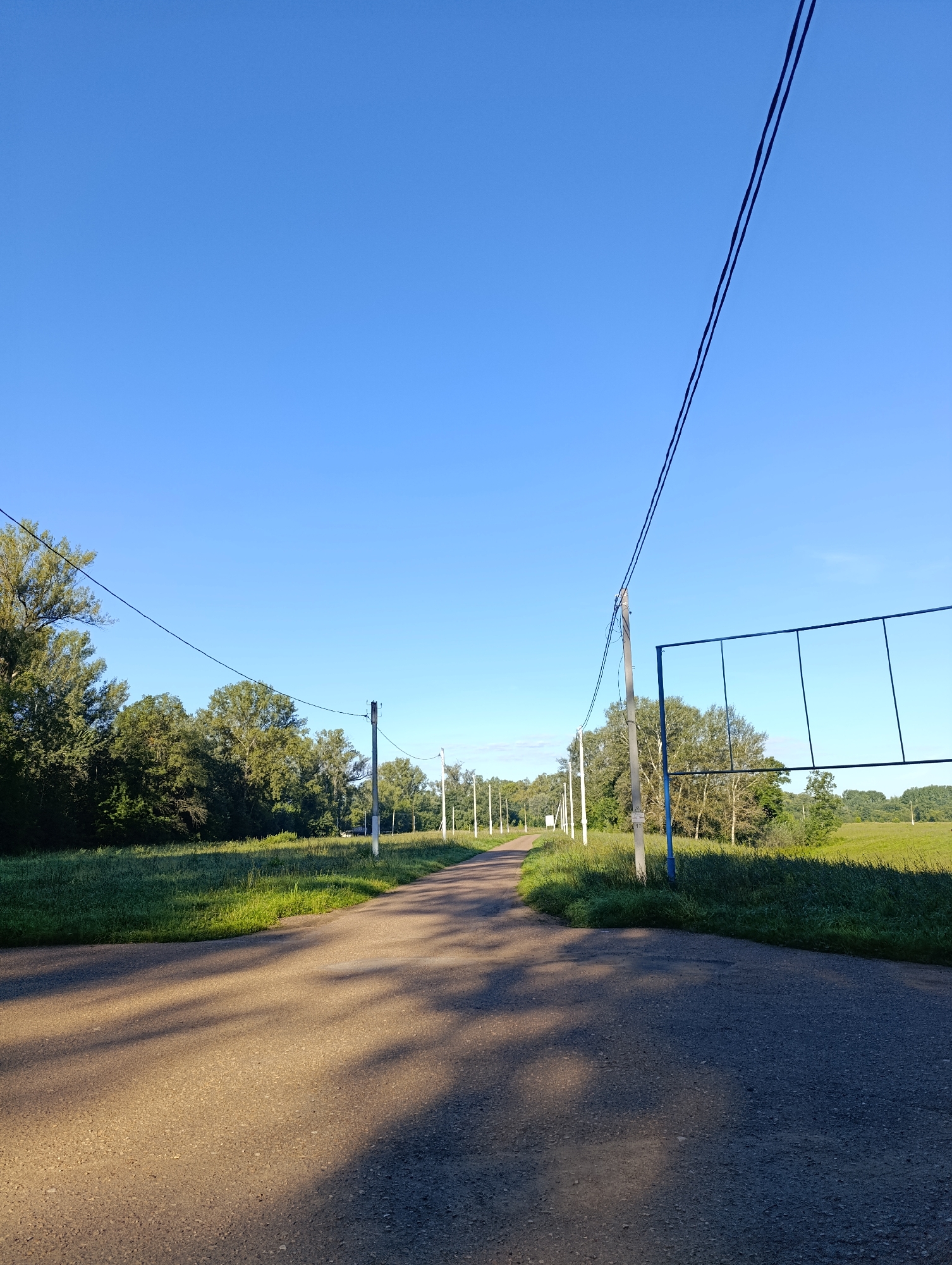 The image shows a paved road in a rural setting. The road curves to the left and is bordered by grassy fields and trees. Overhead power lines run along the road, supported by concrete poles. In the distance, a large rectangular frame with vertical and horizontal bars is visible, potentially part of a sports facility or a billboard. The sky is clear and blue, indicating a sunny day. There are no people or animals visible in the scene. The lighting suggests it is either early morning or late afternoon, given the long shadows cast on the road. The overall atmosphere is peaceful and natural.