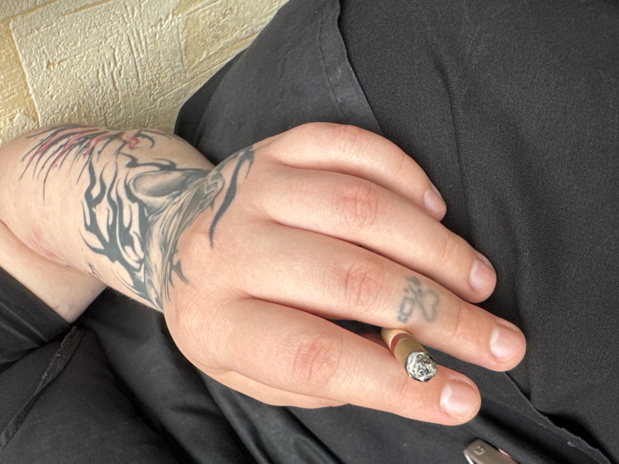 The image shows a close-up of a person's hand and arm, which is heavily tattooed. The person is holding a lit cigarette between their index and middle fingers. The tattoos on the arm appear to be tribal or animalistic in design, with black ink and some red accents. The hand also has a small tattoo on the middle finger, which is difficult to discern clearly but resembles a stylized animal or symbol.
The person is wearing black clothing. The background consists of a textured beige surface, possibly a wall or furniture. The lighting suggests it is daytime, but there are no strong indicators of weather conditions. The scene appears to be indoors due to the controlled lighting and the texture of the background. There is no visible text in the image. The overall impression is candid and unposed, focusing on the details of the tattoos and the act of smoking.