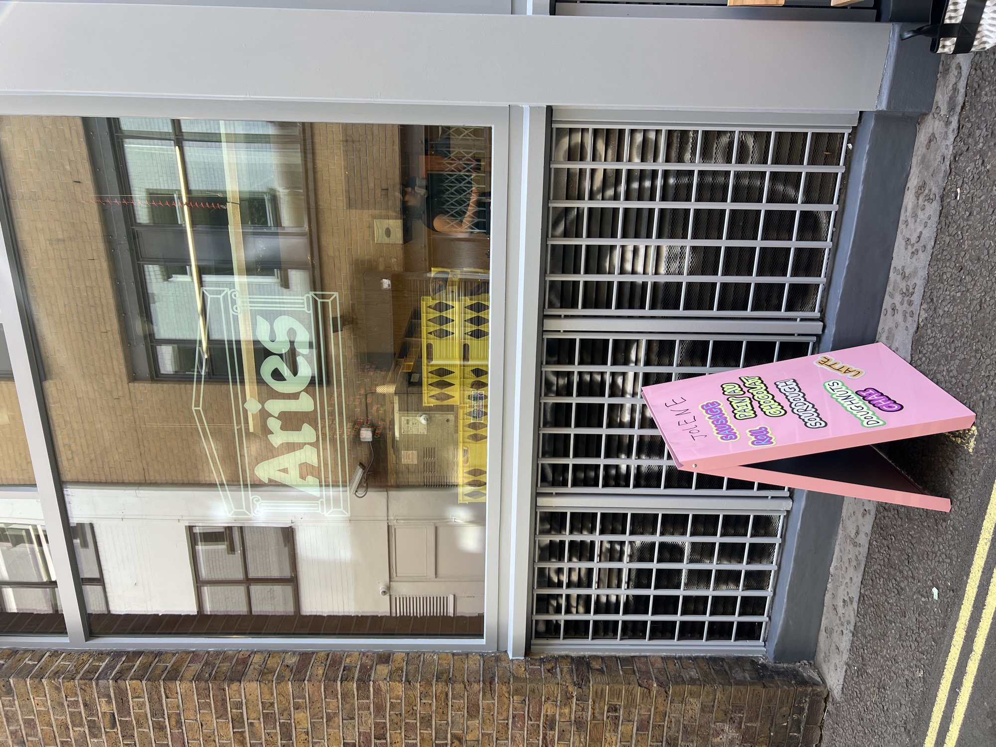 This image captures the exterior of a modern storefront, likely a cafe or bakery, on a sunny day in London, United Kingdom. The left side of the frame features a large glass window, set within a light grey and white frame above a brick wall. Reflected on the window, the business name "Aries" is visible in reverse, indicating it's painted on the inside. Through the glass, reflections of surrounding brick buildings and interior details like yellow storage crates and a security camera are noticeable. A person can be vaguely seen inside the establishment, behind a counter.
To the right of the window are light grey metal grates, possibly covering a basement window or ventilation. On the asphalt pavement in front of these grates, a bright pink A-frame sign advertises various offerings. The pavement also has yellow markings. A small black and white patterned bag is partially visible near the base of the sign.
The text visible on the pink sign, from top to bottom, includes: "DOUGHNUTS", "CHAS" (likely "CHAI"), "LATTE", "Sourdough", "Croissant", "Roll", "Paté Chocolat", "SAUSAGE", and partially visible phrases like "THE V.E." and "TO LEAVE" (possibly "TO TAKE AWAY"). The scene is brightly lit by daylight, suggesting clear weather.