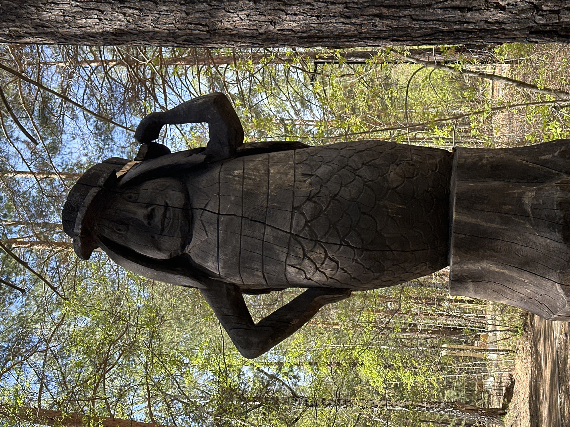 The image shows a wooden sculpture of a human figure with outstretched arms, located in what appears to be a wooded area. The sculpture is dark in color and has a rough, textured surface with some evidence of carving and cracking. The figure is surrounded by trees and foliage. Based on the prompt, the location is Sayansk, Russia.