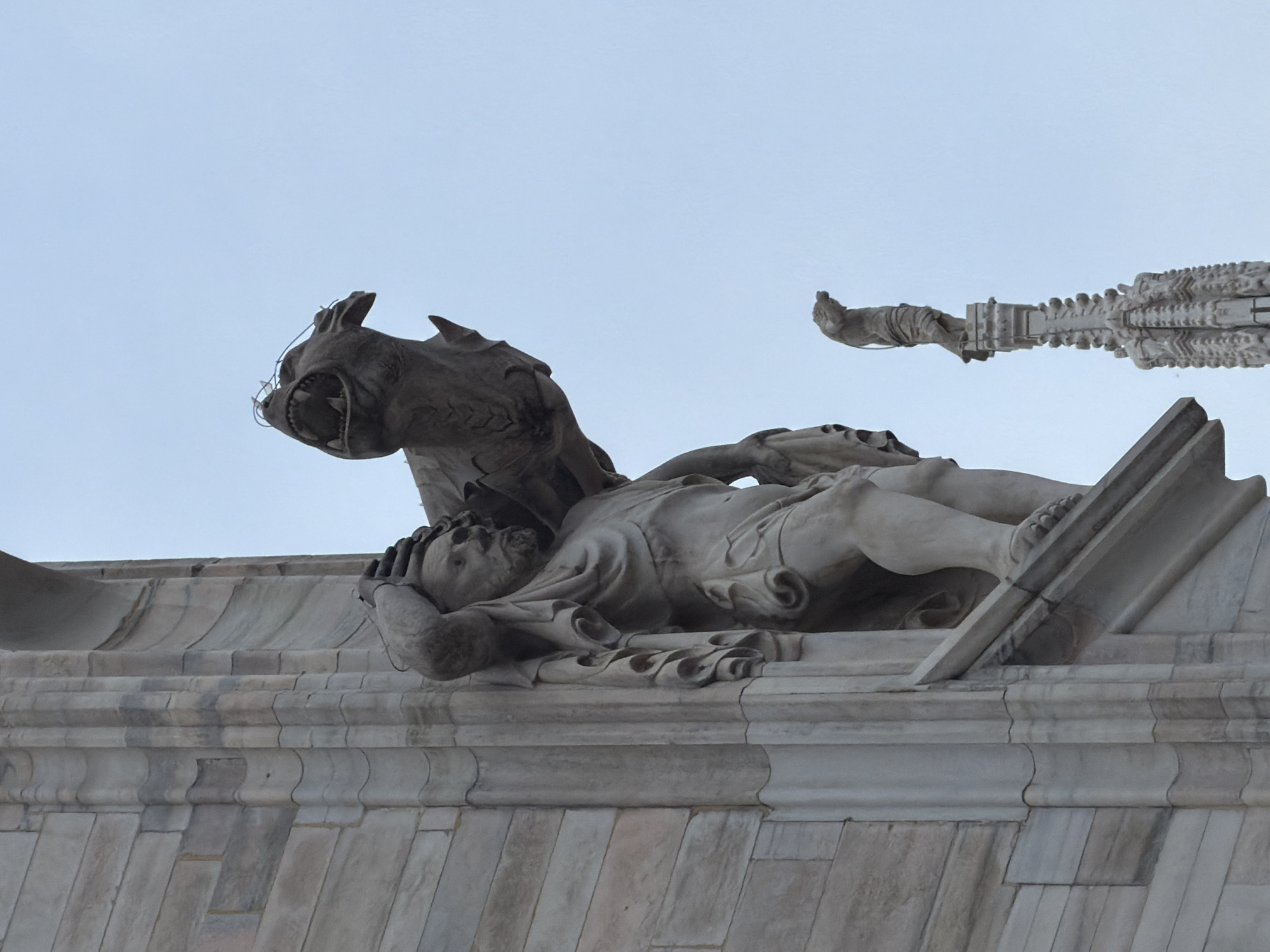 The image captures a dramatic stone sculpture situated on the rooftop of a grand architectural structure, set against a clear, light blue sky. The central sculpture features a human-like figure lying prone, with its hands clasped over its head, conveying an expression of distress or agony. Positioned above and behind this figure is a grotesque, beast-like creature, possibly a gargoyle or demon, with an open mouth revealing sharp teeth, appearing to menace or attack the human. The figures are carved from light-colored stone, likely marble, characteristic of ancient and monumental architecture.
To the far right, a slender, standing human figure is visible atop an intricately carved spiky pinnacle, suggesting another decorative element or small statue. The rooftop itself is composed of finely crafted stone tiles and ornate architectural details, with visible seams and weathering that indicate age. The scene is brightly lit by natural daylight, without harsh shadows, implying fair weather. There is no visible text in the image. The intricate Gothic style and the presence of numerous detailed sculptures strongly suggest this is a view from the rooftop of the Duomo di Milano (Milan Cathedral) in Milan, Italy, known for its vast collection of statues and spires.