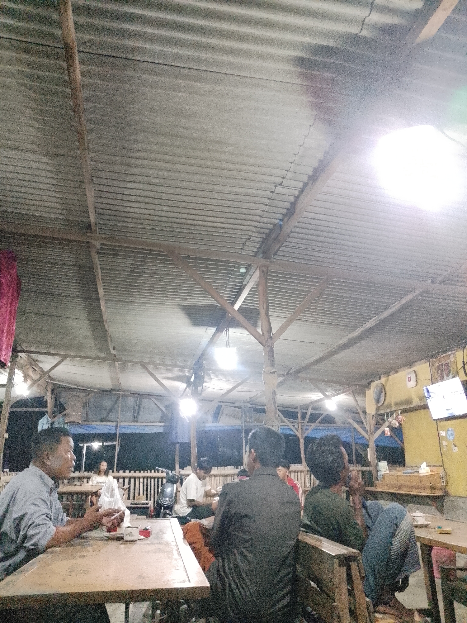 The image depicts an outdoor seating area, likely a warung or informal eatery, under a corrugated metal roof supported by wooden beams. Several people are present, seated at wooden tables.
In the foreground, a man in a grey shirt is seated at a table, holding a cigarette and speaking. On the table in front of him are cups and a pack of cigarettes. To his right, a man in a dark shirt is seated, facing away from the camera. Further back, other individuals are visible, some seated at tables and others possibly walking. A scooter is parked in the background.
The environment appears to be dimly lit, with artificial lights illuminating the space. The corrugated metal roof is dusty and shows signs of age. In the background, a television screen is mounted on a yellow wall. The overall impression is a casual gathering place, possibly in the city of Sugihwaras, Indonesia, given the context provided.
