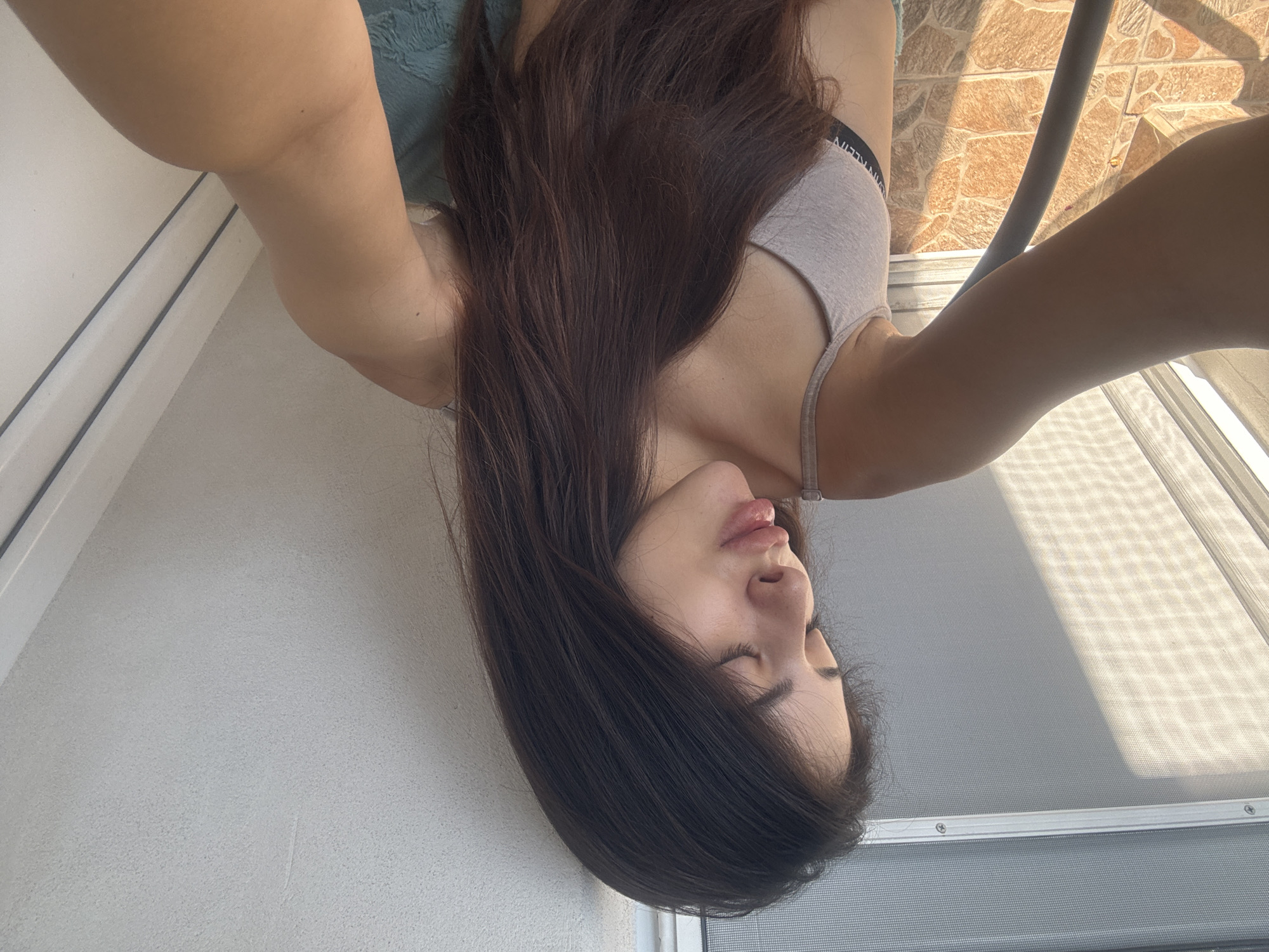 A young woman with long, dark brown hair and closed eyes is positioned in the foreground of the image, looking downwards. She is wearing a light-colored bra with the brand name "CALVIN KLEIN" visible on the band. Her lips are slightly parted, giving the impression of a kiss or pout. Sunlight casts a diagonal shadow across a textured screen or blind in the upper left portion of the image. The background consists of a textured wall and a tiled surface. The woman's left arm is extended towards the viewer, partially obscuring the background. The setting appears to be an outdoor or semi-outdoor location, possibly a balcony or patio. There is no indication of Plovdiv, Bulgaria in the image.