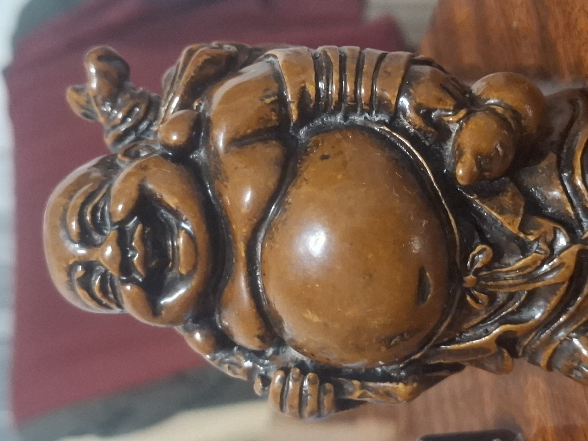 A close-up, eye-level, studio shot focuses on a small, intricately carved wooden figurine of a laughing Buddha, or Budai. The figurine is a warm brown color with dark, carved details highlighting its features. The Buddha has a very large, round belly, a wide, open-mouthed smile, and closed eyes, suggesting mirth. It is draped in flowing robes, tied with a sash at the waist. A sack is slung over its left shoulder, and its hands are positioned to hold an object, though it is unclear what. The figurine is placed on a polished wooden surface, likely a table or shelf. In the blurred background, a dark red fabric or curtain is visible, along with hints of other objects that are out of focus. The lighting is soft and even, creating subtle highlights on the figurine's rounded surfaces. The image captures the texture and craftsmanship of the wooden statue. The location in Almaty, Kazakhstan is not discernible from the image.