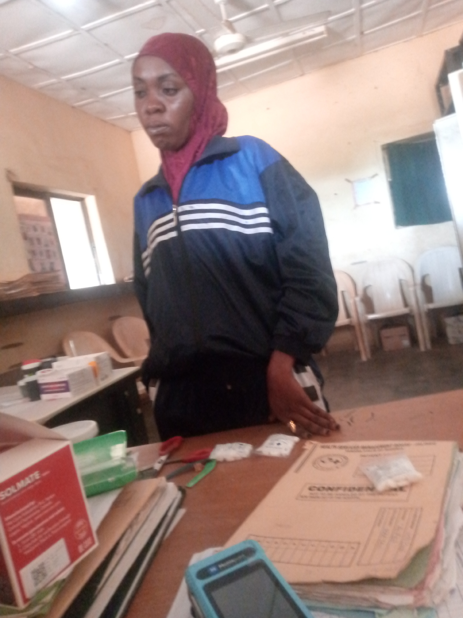 A woman wearing a burgundy headscarf and a blue and black track jacket stands in an office. She is looking down and to her left. The office appears to be dimly lit with fluorescent lighting from a tiled ceiling. In the foreground, a wooden desk is covered with various items, including stacks of papers, a red box labeled "SOLMATE", a green box, scissors, and several small plastic bags containing white pills. In the background, there are chairs, a window with blinds, and a bulletin board on the wall.