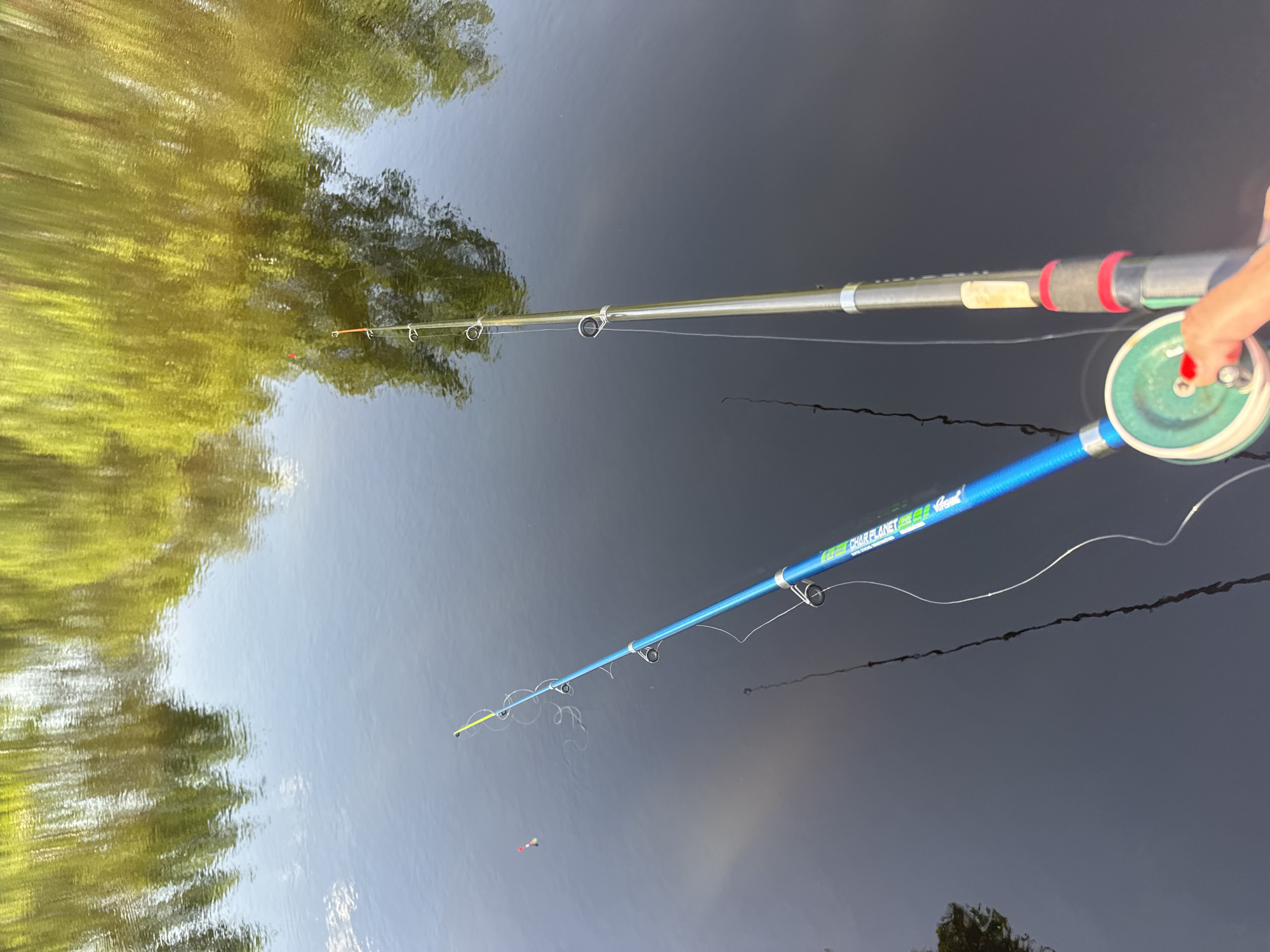 Two fishing rods are extended over a body of water, likely a lake or river, with reflections of trees visible in the water's surface. One rod is silver with a red and black handle, held by a hand with red nail polish, and has a green reel. The other rod is blue with white writing, and fishing line can be seen extending into the water with a red and white bobber attached. The location is not identifiable as Slobodka, Russia.